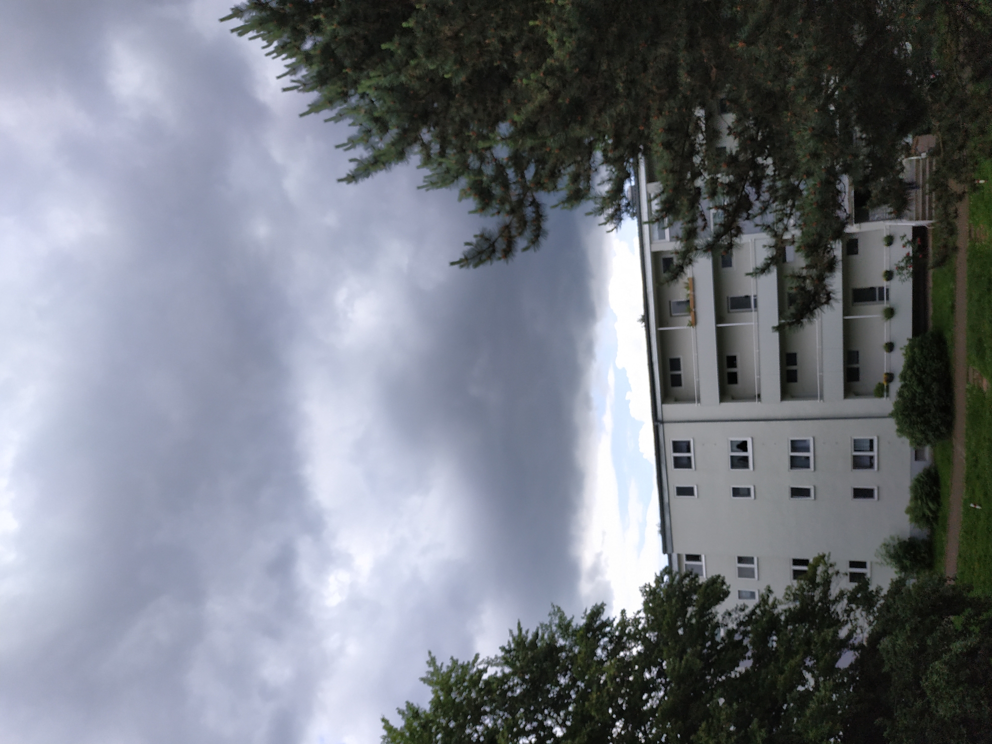 The image captures a residential building under a heavily overcast sky, with lush tree foliage framing the scene. A light-colored, multi-story building, likely an apartment complex, stands prominently, featuring numerous rectangular windows and balconies. Dark green trees, appearing to be a mix of coniferous and deciduous types, occupy the foreground and edges of the frame, partially obscuring the architecture.
The sky is dominated by thick, dark grey clouds, suggesting gloomy, overcast weather, possibly indicating an approaching storm or rain. A small sliver of lighter sky can be seen on the right, contrasting with the darker cloud mass. On the ground level to the right of the building, a strip of green grass and a pathway are visible.
No people or specific activities are taking place in the scene, which appears static and devoid of human presence. There is no visible text. The overall impression is one of impending weather, with the heavy cloud cover creating a somber or contemplative atmosphere.