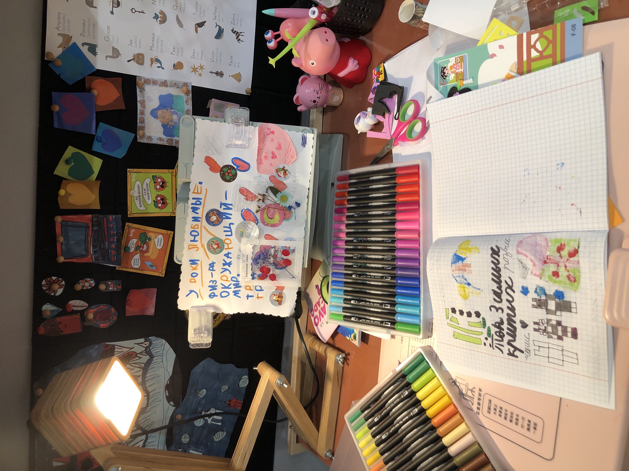 The image shows a desk with various art supplies and decorations. A lamp is on, illuminating the scene. Several papers with drawings and writing are pinned to a dark background. On the desk, there are markers, scissors, a notebook with drawings, and some figurines. The notebook has some handwritten text in Russian, along with drawings of what appear to be cartoon characters and abstract shapes. There is also a paper with a list of words in English and Russian, possibly for learning purposes. The overall impression is of a child's creative workspace. No specific location in Saint Petersburg, Russia can be determined from the image.