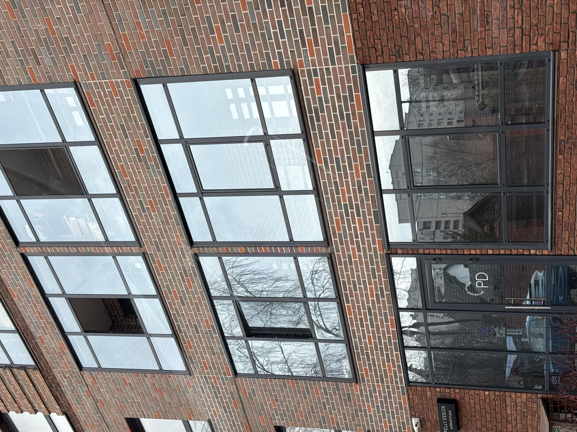 The image shows the exterior of a brick building with large, multi-paned windows. The building facade is predominantly red brick with sections of gray brick. Some of the windows are open. The windows reflect the surrounding environment, including a building across the street and bare tree branches. The building appears to be in a commercial or office district. A sign on the building reads "POLLY DESIGN INTERIOR STUDIO". A logo with the letters "PD" is visible on one of the windows.
The location in the city of Izhevsk, Russia, is specified.