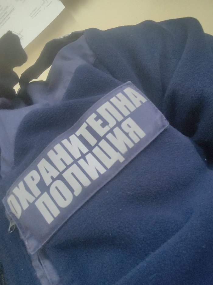 A dark blue fleece uniform jacket with lighter blue fabric accents is depicted from a slightly overhead perspective. A rectangular light blue patch, affixed to the jacket, displays white capitalized Cyrillic text that reads "ОХРАНИТЕЛНА ПОЛИЦИЯ" (Security Police). The garment rests on an unidentifiable light-colored surface. In the upper left background, a portion of a white document with printed text and a partial handwritten signature is visible. No individuals or active interactions are present in the frame. The scene is located in the city of Unknown, Unknown.