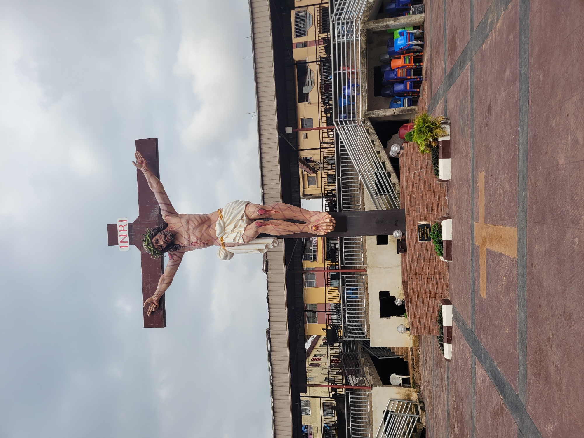 The image shows a life-size statue of Jesus Christ on a cross, set in an outdoor paved area. The statue depicts Jesus with a crown of thorns, a loincloth, and wounds on his body. The cross is large and brown, with the letters "INRI" at the top. Behind the statue, there is a building with a corrugated metal roof, balconies, and multiple stories, appearing to be residential or commercial units. Steps and railings lead up to the building, and a stack of colorful plastic chairs is visible under a covered area. The ground is paved with reddish-brown tiles and features a cross-shaped pattern. The sky is overcast with gray clouds. A plaque is visible at the base of the statue. This scene is located in Onitsha, Nigeria, and is likely part of a church or religious site.