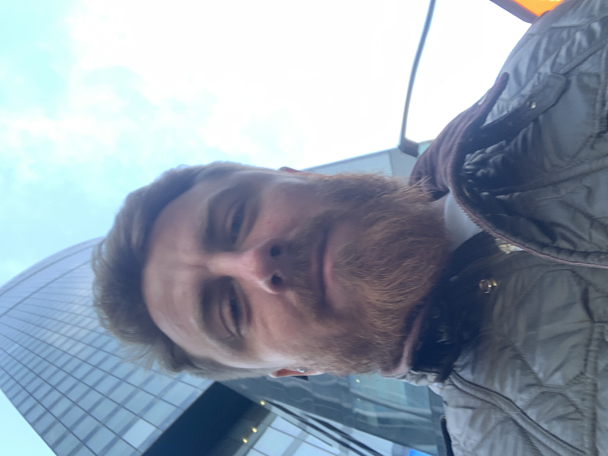 A selfie taken from a low angle features a man with light skin, light brown hair, and a ginger beard. He is wearing a quilted jacket, possibly grey or olive green, with a brown collar. Behind him, a tall, modern building with a glass facade and a grid-like pattern reflects the cloudy sky. The building's design suggests a location in a modern city. The location in the city of Khimki, Russia cannot be confirmed.