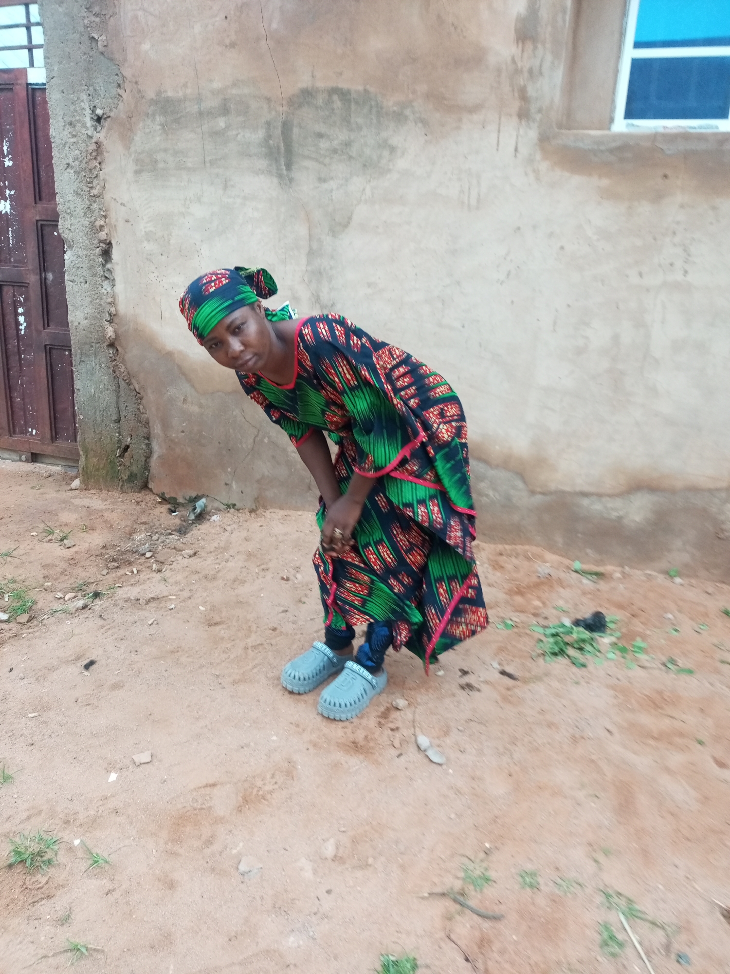 A woman wearing a green and black patterned dress with pink trim and a matching headscarf is bending over in a sandy, outdoor area in front of a concrete building. Her feet are clad in light gray slip-on shoes. The building has a weathered, textured wall and a window with a blue pane. A dark brown door with vertical planks is visible to the left. The ground is covered in sand and scattered small stones and patches of green vegetation.