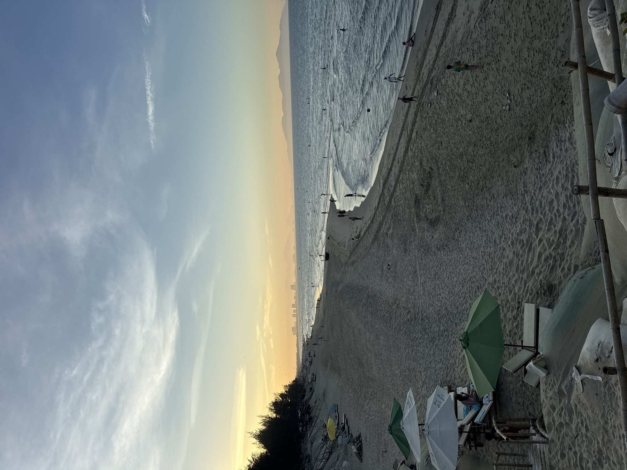 The image captures a beach scene. The sky transitions from a light blue to a yellow-orange hue towards the horizon. The sea shows waves near the shore, with people visible in the water and on the sand. The beach itself has a sandy texture, marked with footprints and the presence of beach umbrellas and lounge chairs. A line of trees or vegetation runs along part of the shoreline. Further in the distance, a silhouette of a city skyline is visible along the horizon, with mountains beyond.