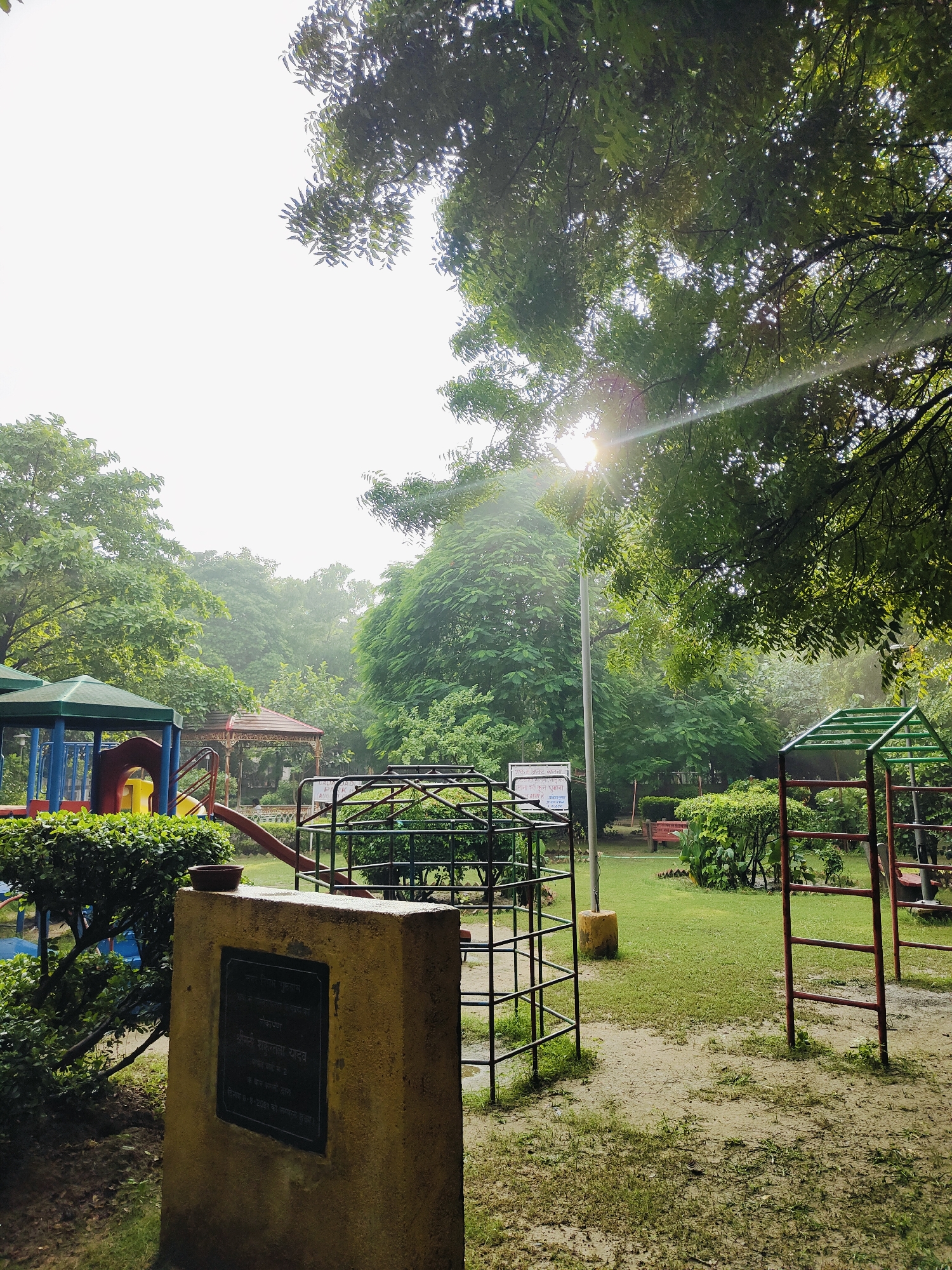 The image depicts a children's playground in Gurgaon, India. In the foreground, a yellow concrete pillar with a dark plaque is visible. The plaque appears to contain text in Hindi, possibly an inscription or memorial. To the left of the pillar, a green bush and part of a colorful playground structure with a blue and green roof and red and yellow slides can be seen. A metal climbing frame is also present in the center of the image. In the background, more playground equipment, trees, and lush green grass are visible. The sun is shining through the trees, creating a lens flare effect. The overall environment suggests an outdoor recreational area.