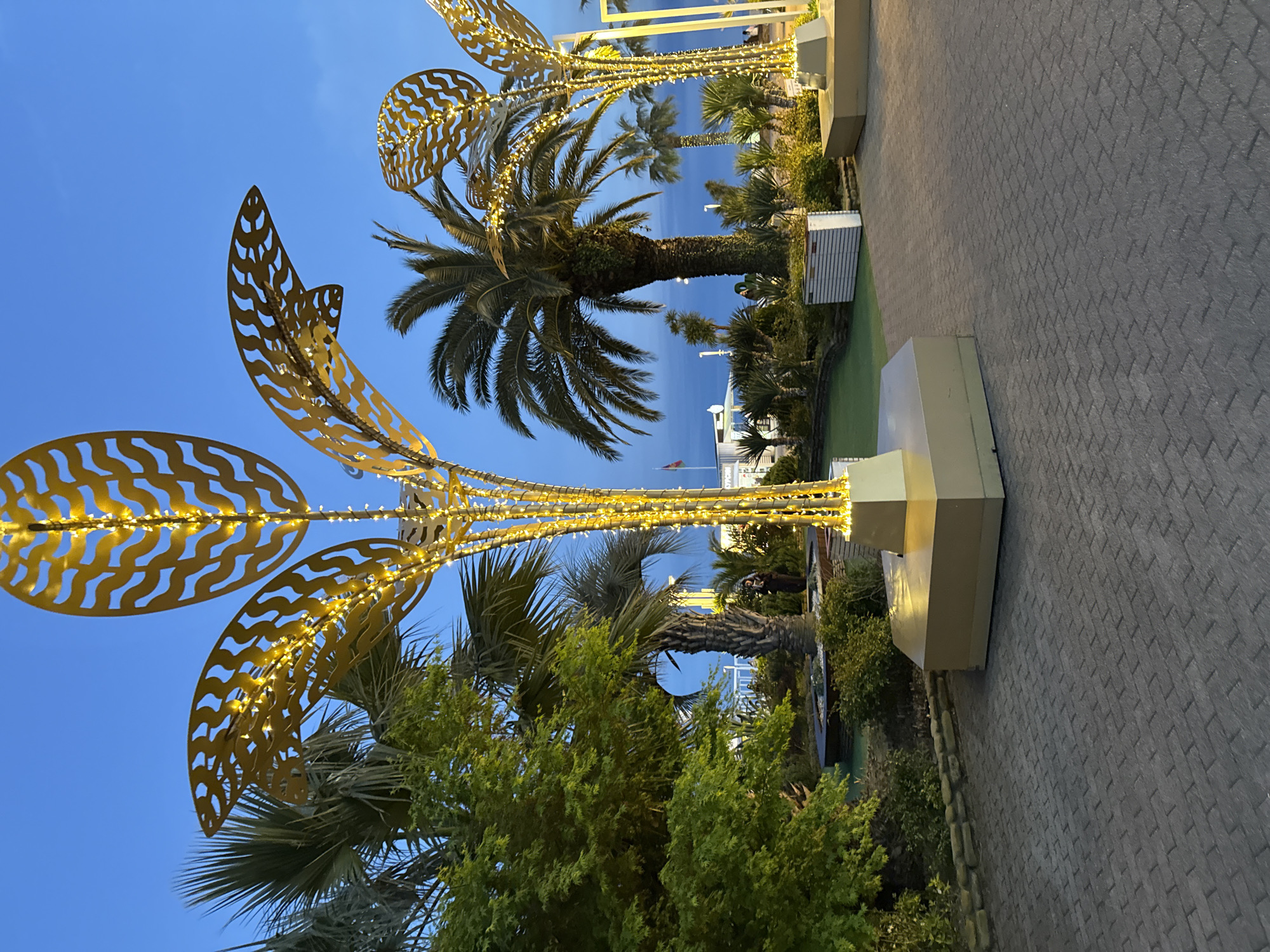 The image, taken at twilight or early evening, showcases an outdoor promenade or landscaped area. In the foreground, large, golden, ornate structures, designed to resemble stylized palm fronds or leaves, are brightly illuminated with numerous small, warm-toned lights. These decorative elements suggest artificial trees or festive lighting fixtures.
Beneath and around these golden structures, several natural palm trees with thick trunks and lush green fronds are visible, intermingled with various green bushes and other low-lying foliage. The ground features a wide paved walkway made of dark gray interlocking pavers, stretching across the right side of the frame. Along this walkway are light-colored, rectangular structures, possibly benches or planters, some also subtly lit.
The sky is a clear, deep blue, indicative of dusk or dawn, with no clouds. In the background, beyond the trees and landscaping, a body of water (likely a sea or large bay) is visible, with distant buildings or structures along the shoreline. A small flag on a pole can be discerned in the far distance. The scene is static, devoid of people or specific activity, and evokes a peaceful, inviting, and somewhat festive atmosphere typical of a resort or coastal public space. No discernible text is present.