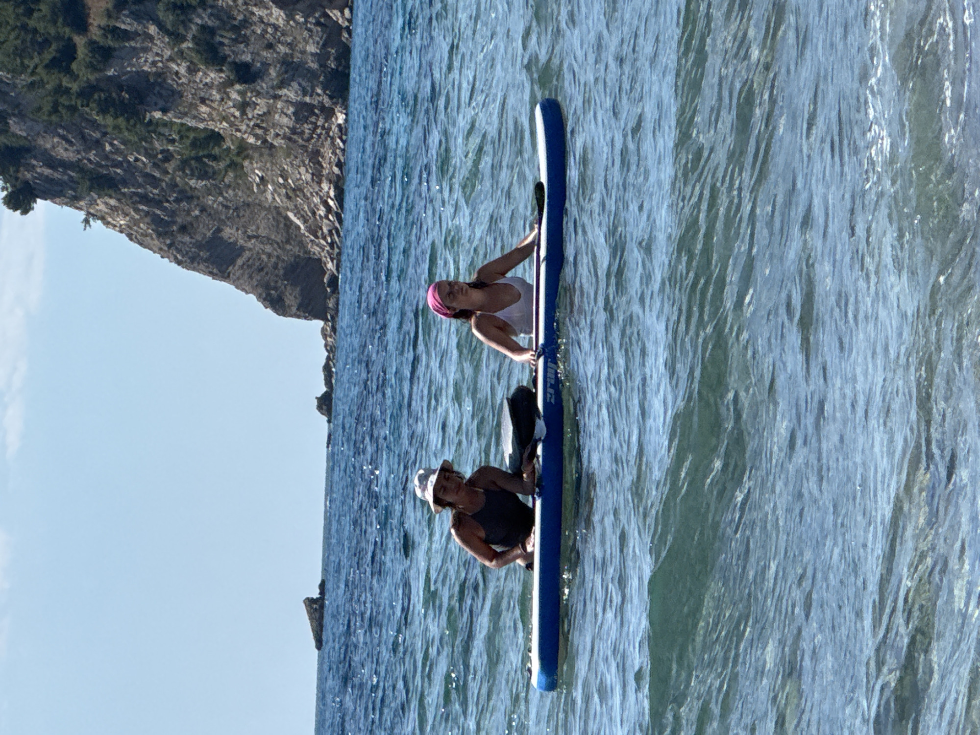 The image shows two women on paddleboards in the sea. The woman on the left is wearing a dark swimsuit and a light-colored hat. She is lying on her stomach on the paddleboard, with her arms resting on it. The woman on the right is wearing a white swimsuit and a pink bandana. She is sitting on the paddleboard, holding a paddle. The paddleboards are blue and white. In the background, there is a rocky cliff with green vegetation and the sea. The sky is clear and blue. The location is the coast of Đurovići, Montenegro.