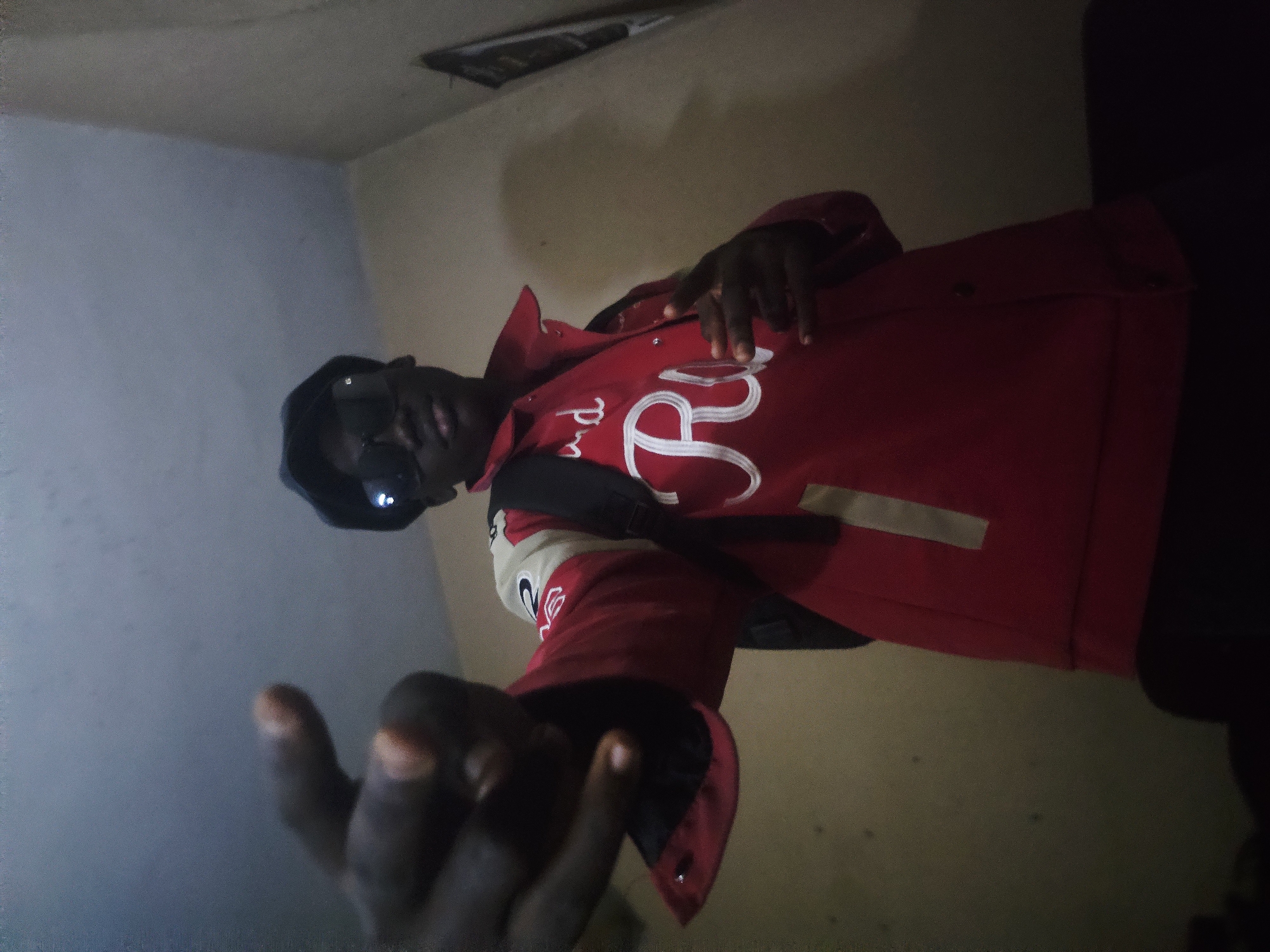 A person is posing for a photo indoors. The individual is wearing a red jacket with white lettering on the back, sunglasses, and a cap. The setting appears to be a dimly lit room with cream-colored walls. A backpack is slung over the person's shoulder. The photo was possibly taken in Nkwelle, Nigeria, as per the prompt.