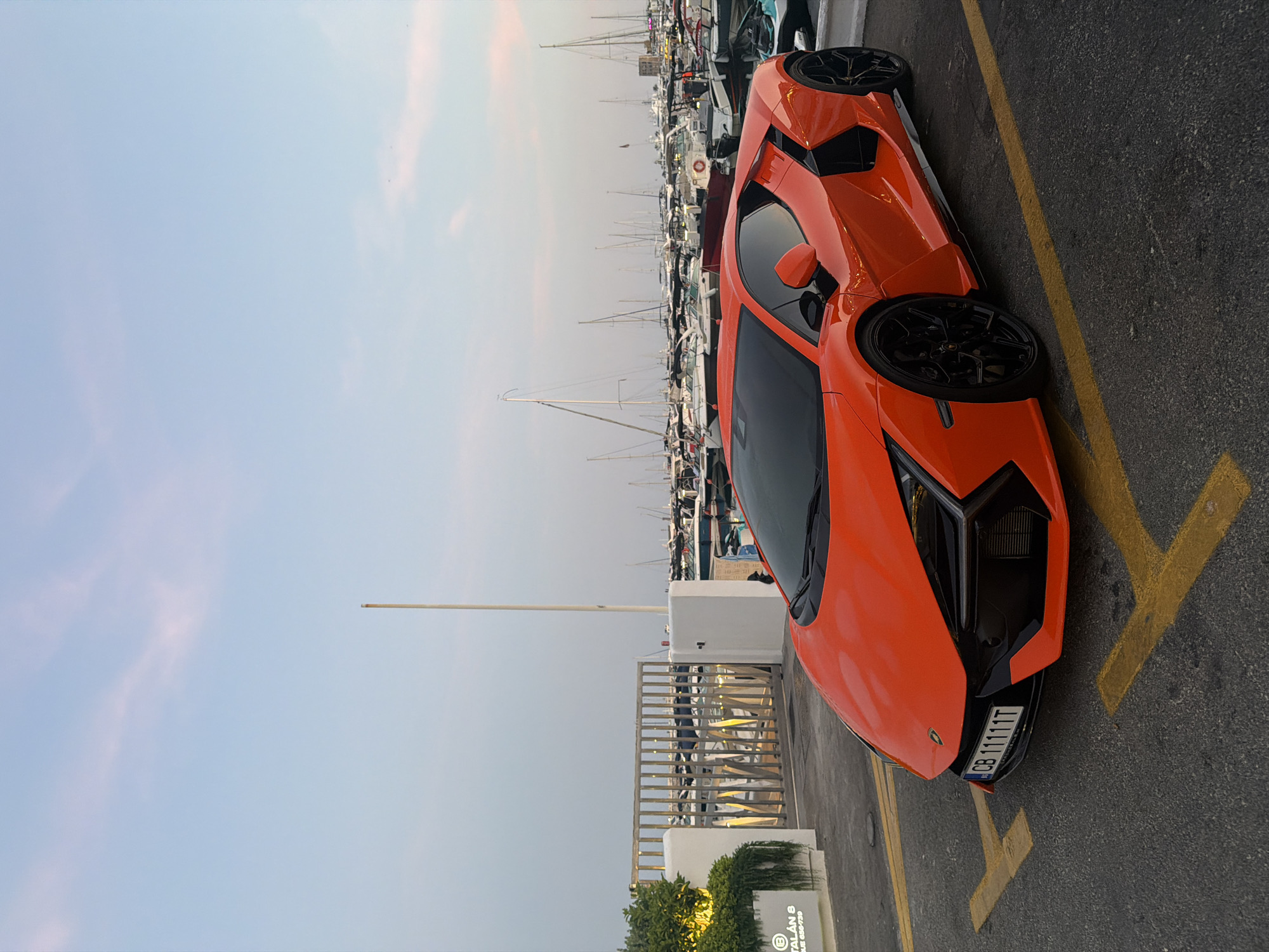 An orange Lamborghini is parked in a parking lot adjacent to a marina filled with yachts. The sky above is a soft blue with light pink clouds. The car is positioned in the foreground, facing left, with its front bumper and hood clearly visible. The license plate reads "CB 11111T". In the background, a white building with a metal gate and a sign reading "CATALAN B" is visible. Beyond the gate, numerous sailboats and motorboats are docked in the marina. The ground is asphalt with yellow parking lines.
