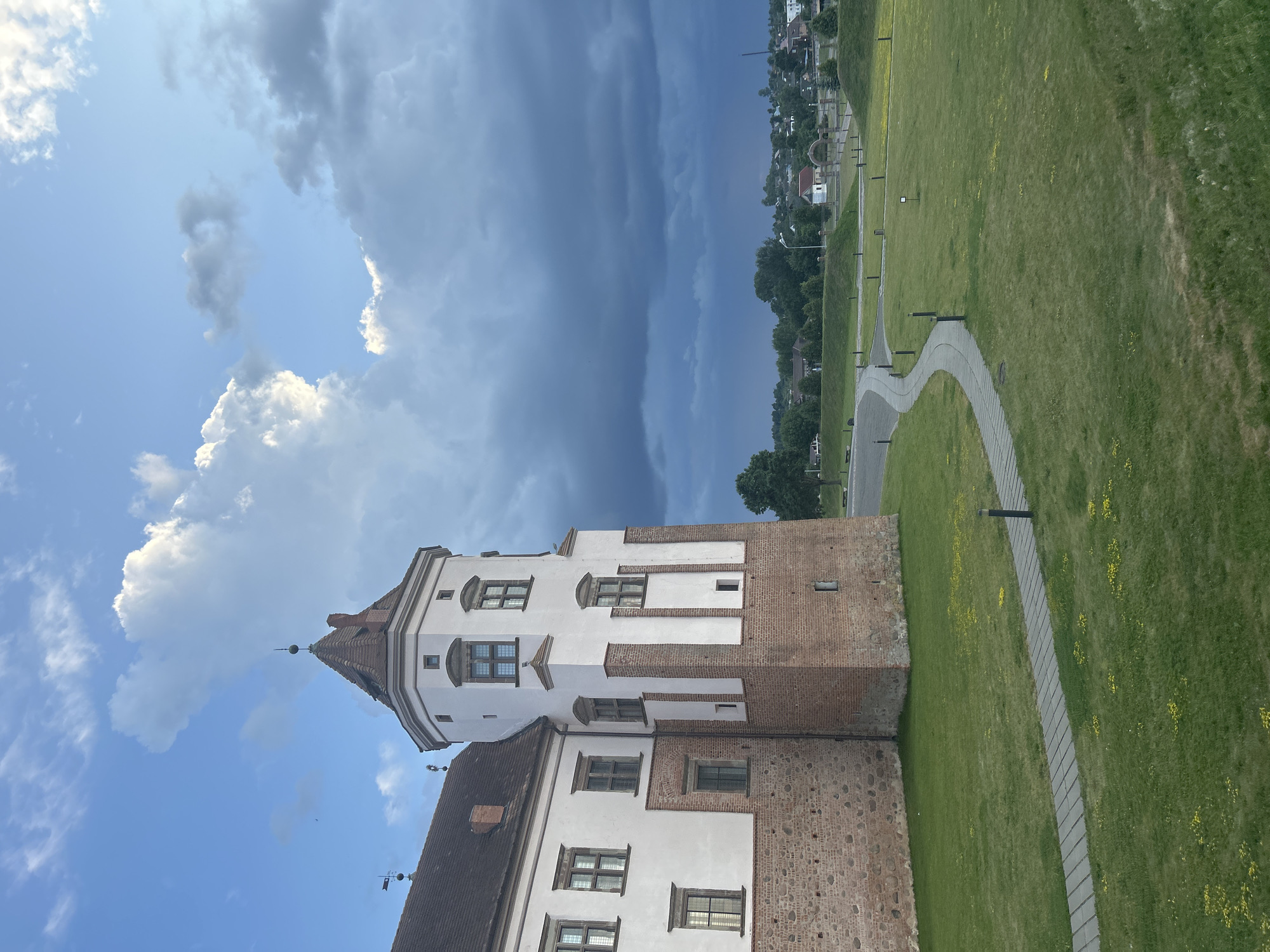 The image depicts a view of the Mir Castle Complex in the village of Mir, located within the Zhukhavitski Sel'ski Savet, Belarus. The foreground features a neatly mown green lawn with a paved pathway winding through it. Small yellow flowers are scattered across the grass. The pathway is illuminated by several dark, cylindrical outdoor lamps. To the left, a section of the Mir Castle is visible, characterized by its red brick base and white plastered upper walls. A prominent white tower with a red tiled roof and a weather vane stands prominently on the castle's corner. Numerous windows with dark frames are visible on the castle walls. In the background, a rolling landscape with trees and buildings can be seen under a dramatic sky filled with both bright white clouds and darker, stormier clouds. The overall scene suggests a late afternoon or early evening setting with approaching weather.