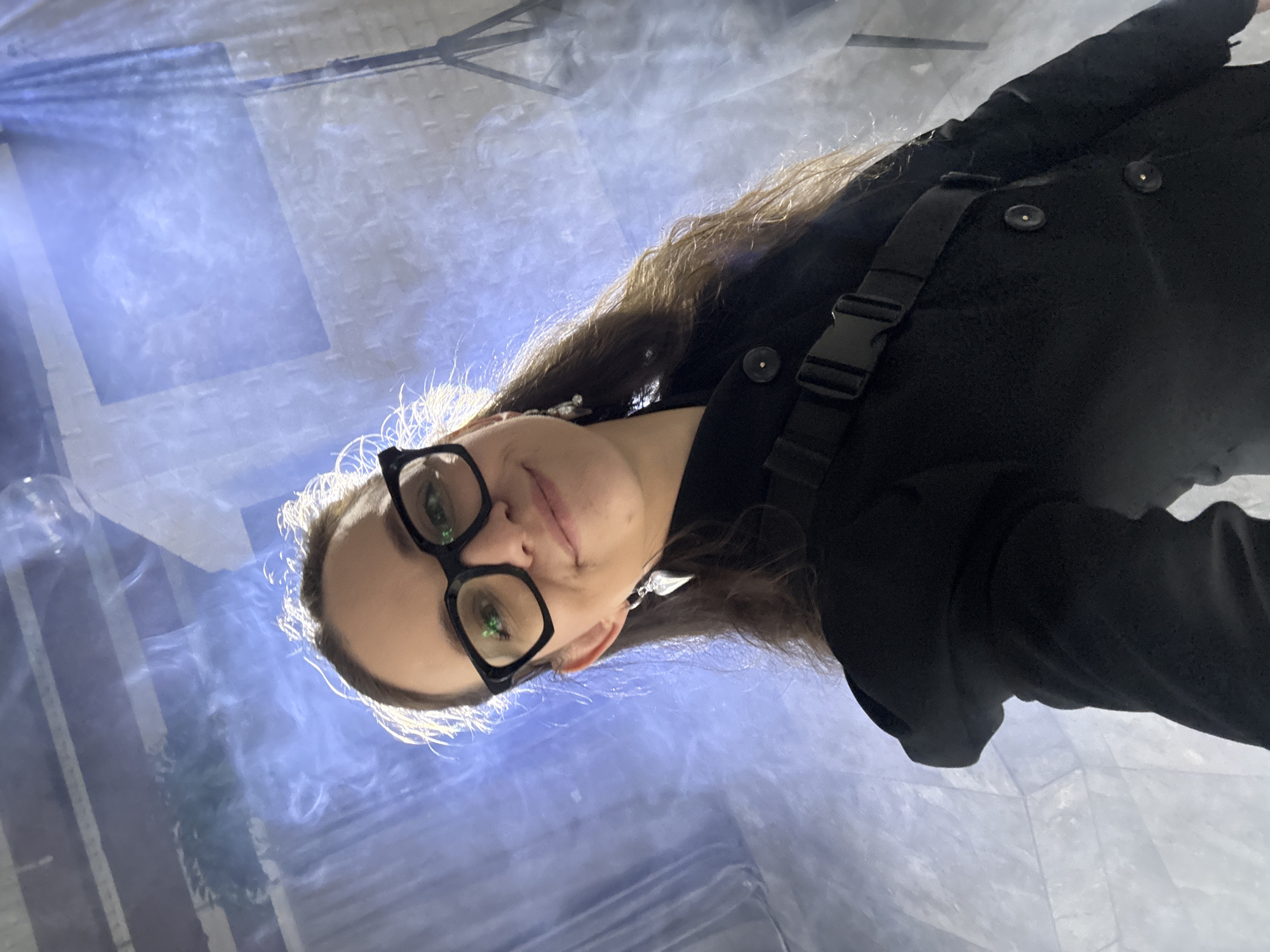 A young woman with long, dark blonde hair and glasses is shown from a low-angle perspective, looking upwards. She is wearing a black jacket with a buckle strap across the chest and two visible buttons. The background is a bright, textured surface with blue and white hues, possibly a wall or a large sheet of fabric, with streaks of light and mist. She appears to be indoors. The lighting suggests the photo was taken with artificial light, possibly a spotlight or flash. Her expression is neutral, and the overall mood is somewhat artistic or dramatic due to the lighting and angle. There is no visible text.