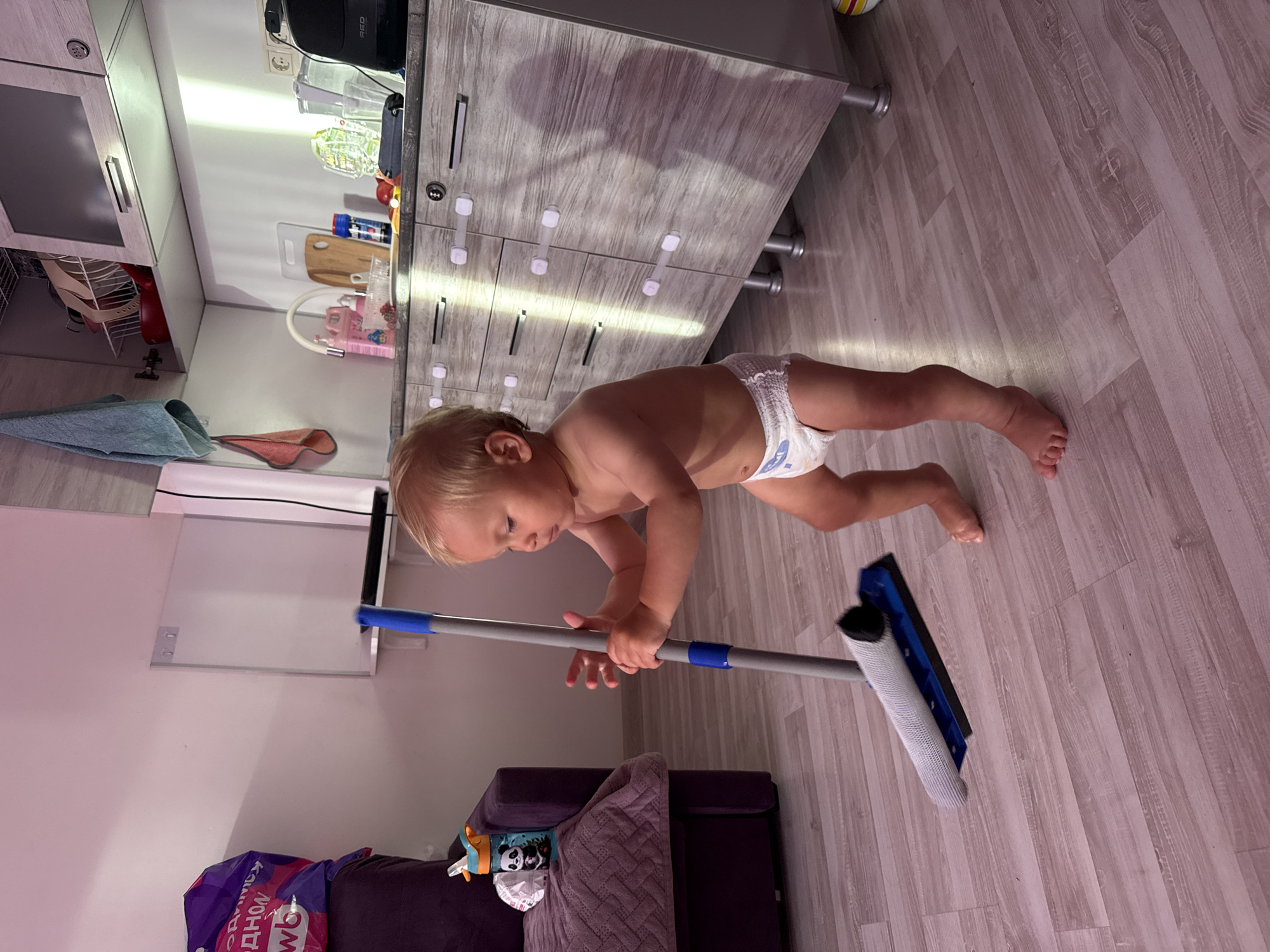 A blonde-haired toddler wearing a diaper is standing on a light-colored wooden floor and holding a mop. The toddler is facing left and looking down at the mop. The mop has a blue handle with blue accents and a gray cleaning head. In the background, there is a kitchen cabinet with light-colored wood grain, kitchen items on the counter, and a purple sofa. The lighting in the room is warm, with sunlight casting shadows on the floor and cabinets. The location is not specified in the image.