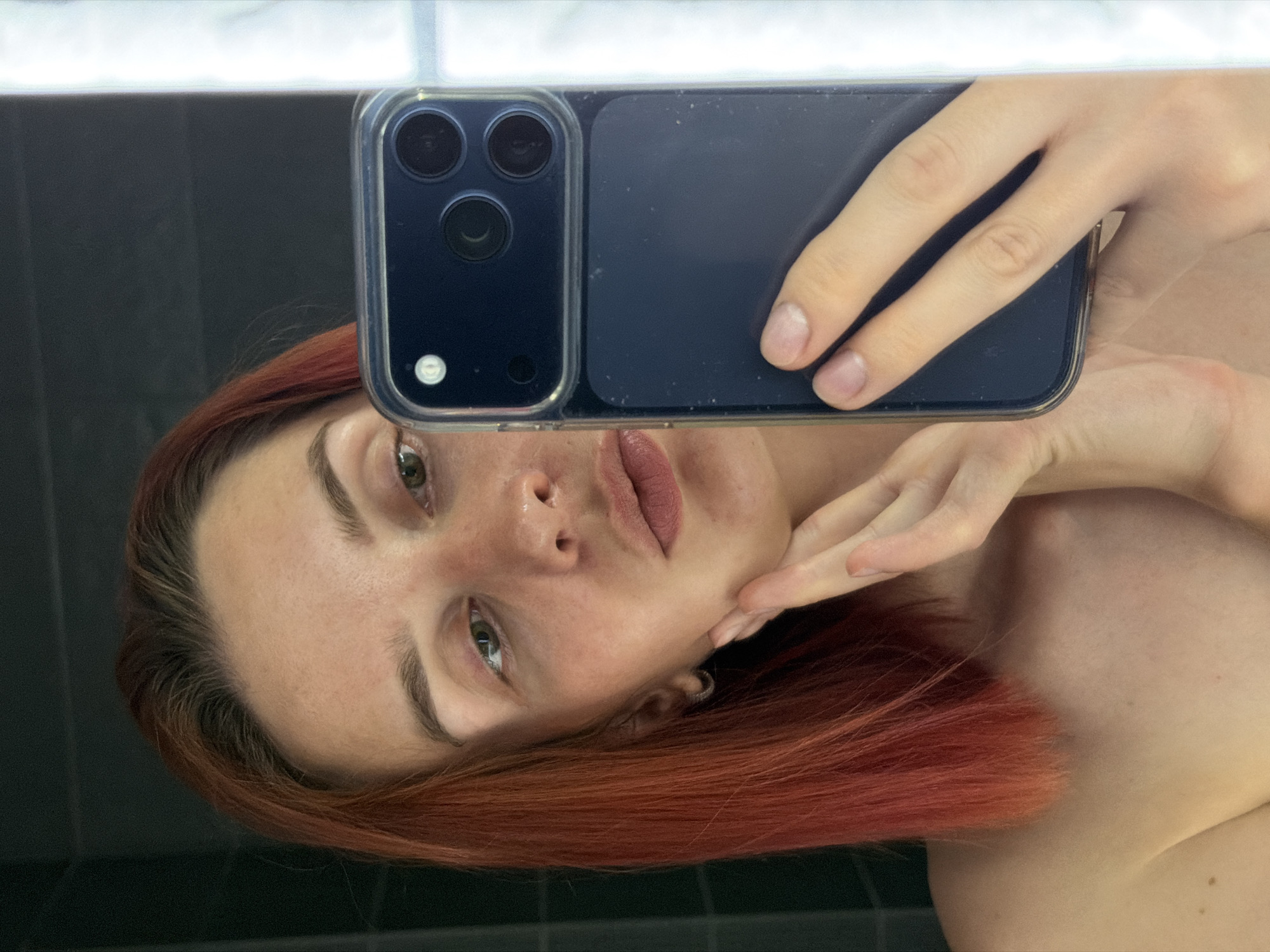 A woman with reddish-brown hair takes a selfie in a bathroom with dark tiling. She is holding a smartphone with a dark blue case in front of her face. The location is unknown, and there is no indication that it is in the city of Chaikovsky, Russia.
