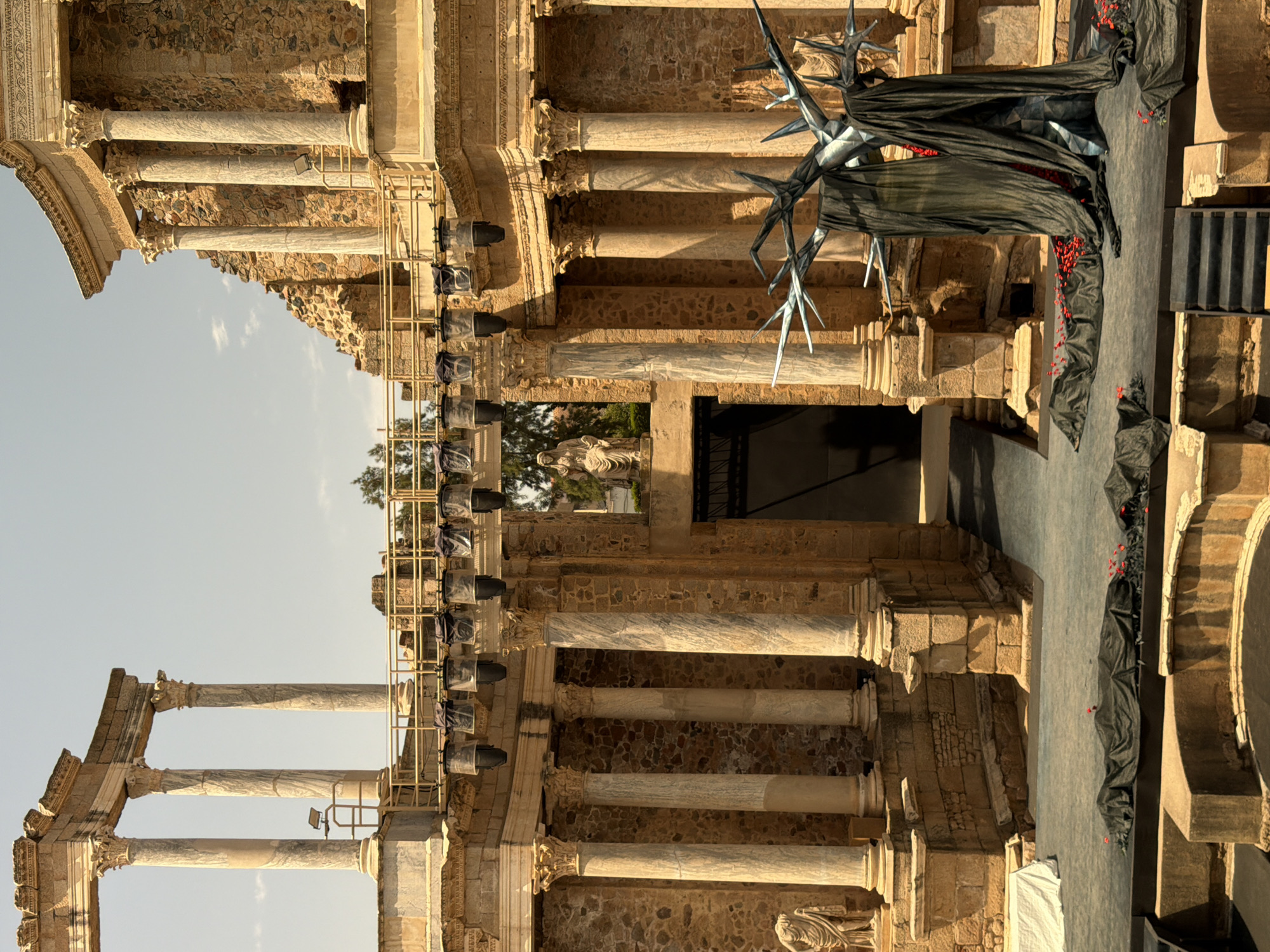 The image depicts the Roman Theatre of Merida, Spain, a UNESCO World Heritage site. The theatre, an ancient stone amphitheater, serves as a stage for a theatrical production. The stage is adorned with dark fabric draped over a metallic tree-like sculpture with sharp, angular branches. Scattered red rose petals are strewn across the stage floor, some nestled within the fabric. In the background, the stone seating of the theatre rises, interspersed with imposing columns and remnants of the ancient structure. The upper portion of the theatre reveals a clear sky, suggesting daytime. A few statues are visible, partially integrated into the architectural features. Stage lighting is suspended from a metal rig above the stage, hinting at an upcoming or concluded performance.