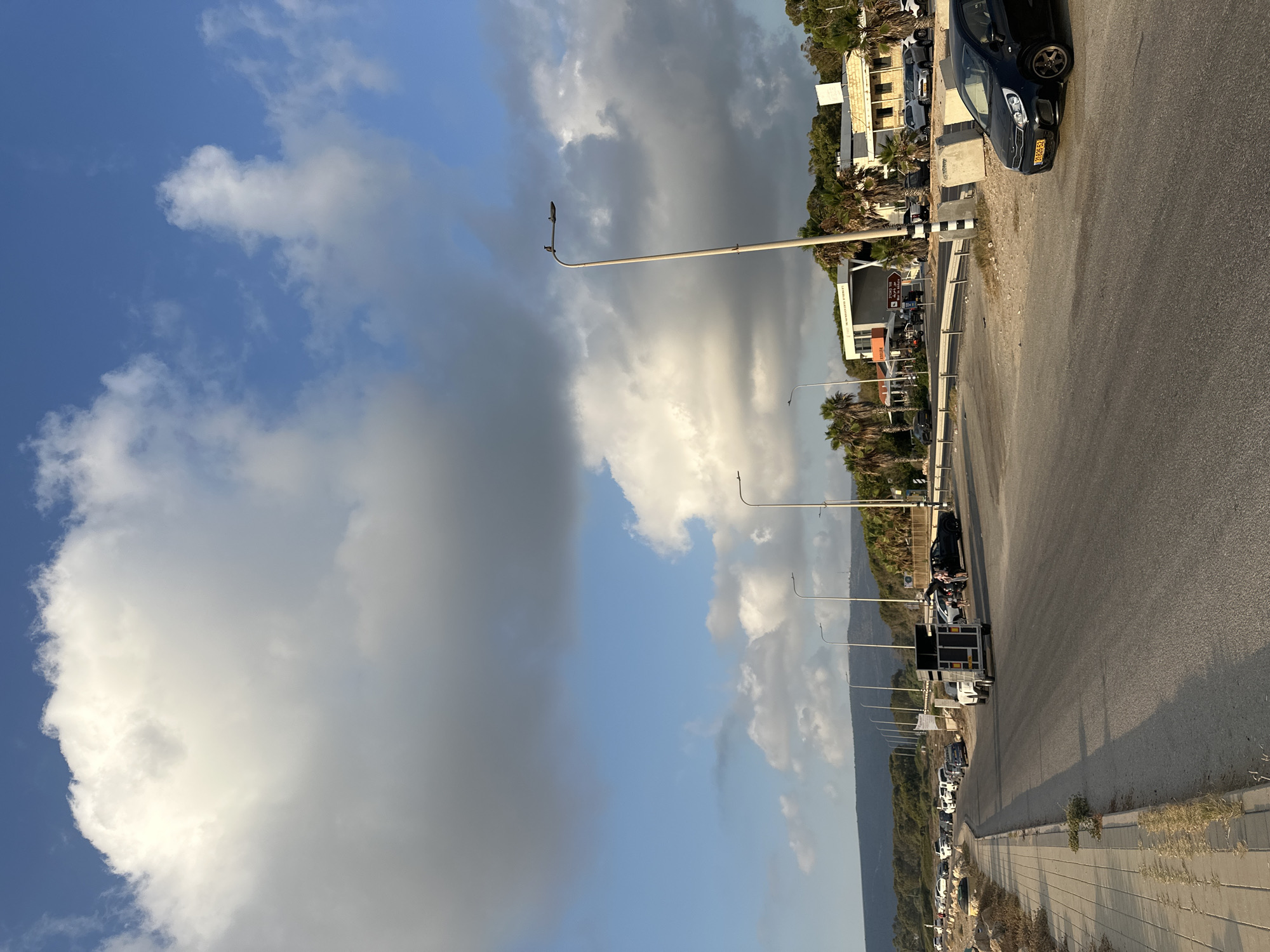 A street scene in Nahariyya, Israel, shows a paved road with parked vehicles and a sidewalk. The sky is partly cloudy, with large white clouds against a blue background. On the right side of the road, there is a street lamp with a bird perched on it. Several cars are parked along the road, and some people are standing near them. In the background, there are buildings, palm trees, and a hill. The overall scene appears to be a public area with commercial establishments and residential buildings.