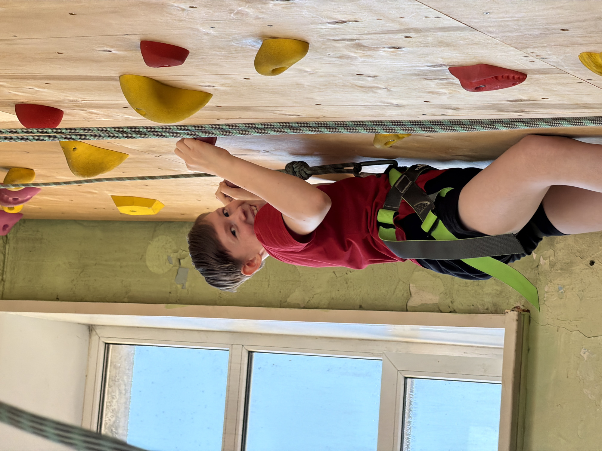 A young boy is actively engaged in indoor rock climbing, depicted in an image rotated 90 degrees counter-clockwise from its upright orientation. He is positioned on a light-colored wooden climbing wall, which is dotted with numerous brightly colored climbing holds in red and yellow, varying in shape and size. The boy, appearing to be in his pre-teen years, has short, dark brown hair and is wearing a red short-sleeved t-shirt, dark shorts, and a neon green and black safety harness. A striped green and black climbing rope is prominent, running horizontally across the wall and also attached to his harness via a carabiner, indicating he is secured. He is holding onto the climbing holds with both hands, his body angled as if in mid-climb or resting.
The boy is looking directly at the camera with a wide, happy smile, conveying enjoyment and confidence. Below the climbing wall, a section of an older light green wall with peeling paint and cracks is visible, suggesting an established, possibly aged, indoor facility. A white horizontal ledge or window sill separates this lower wall from a large window with white frames. Through the window, a clear, bright blue sky is visible, indicating daytime. The scene takes place indoors, likely at a climbing gym or sports facility in Izhevsk, Russia. There is no visible text in the image.