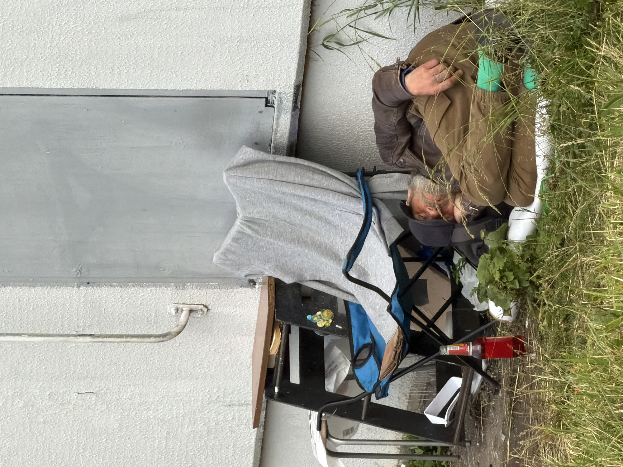 A man sleeps outdoors in front of a building, leaning against a folding chair. He is wearing a brown leather jacket, khaki pants, and a dark cap. His head rests on a pillow or cushion on the ground, with his face turned away from the camera. A grey blanket is draped over the chair. Next to the chair, on the ground, is a red bottle of what appears to be a beverage. There is also a small pile of green foliage and a white object next to the man. A black portable table with a wooden top is set up behind the chair, holding some papers and other items. The building wall is white and textured, with a grey metal door to the right of the scene. Green grass and some taller weeds surround the sleeping area. The lighting suggests it is daytime, possibly overcast.
