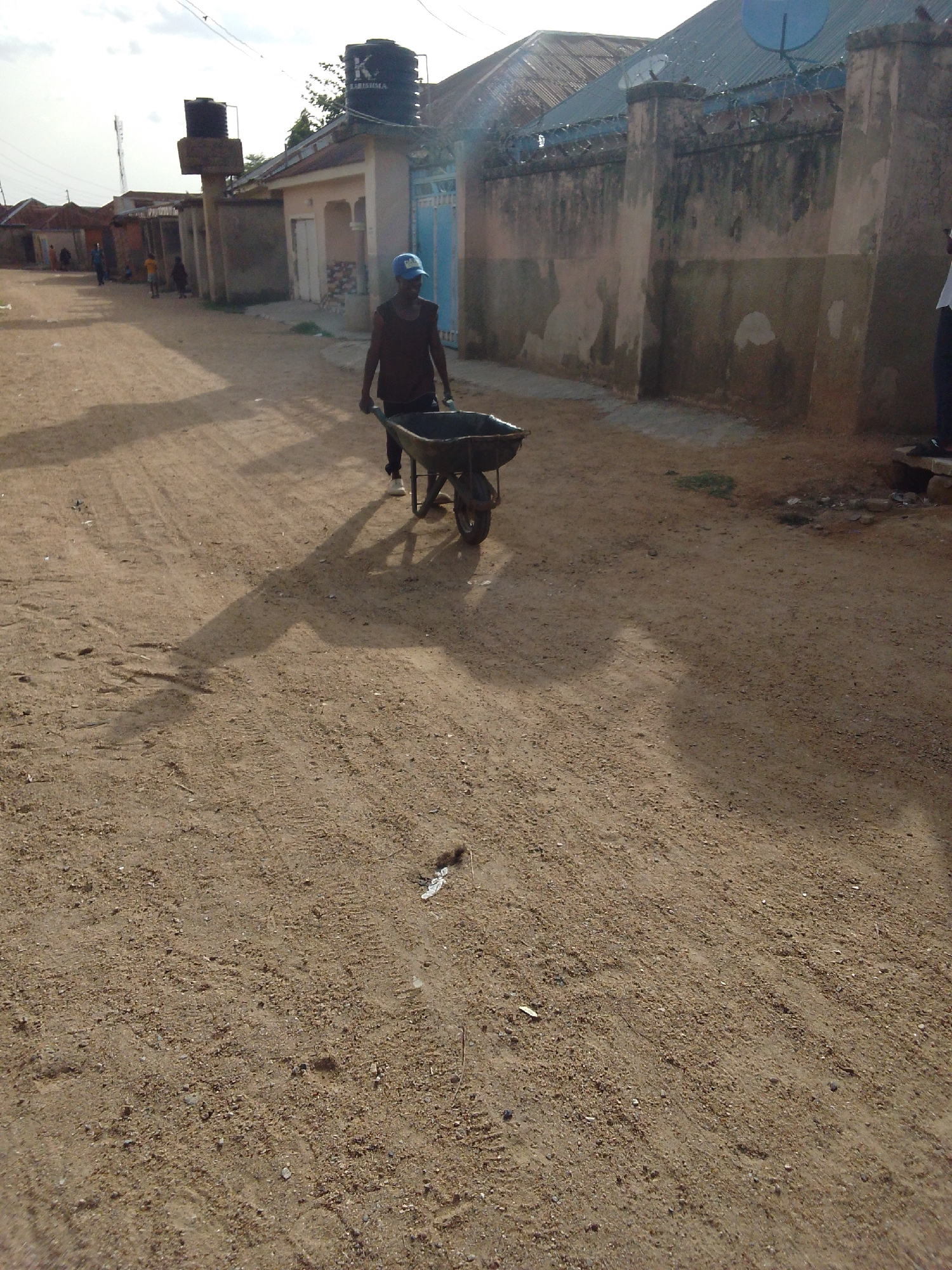 A man is pushing a wheelbarrow down a dirt road in Bauchi, Nigeria. The road is unpaved and dusty, with tire tracks visible in the sand. Buildings and walls line the sides of the road. A blue water tank sits on top of a building in the background. The man is wearing a dark-colored shirt, dark pants, white shoes, and a blue cap. The wheelbarrow is dark-colored and appears to be empty. The scene is bathed in bright sunlight, casting long shadows. A satellite dish is visible in the background.