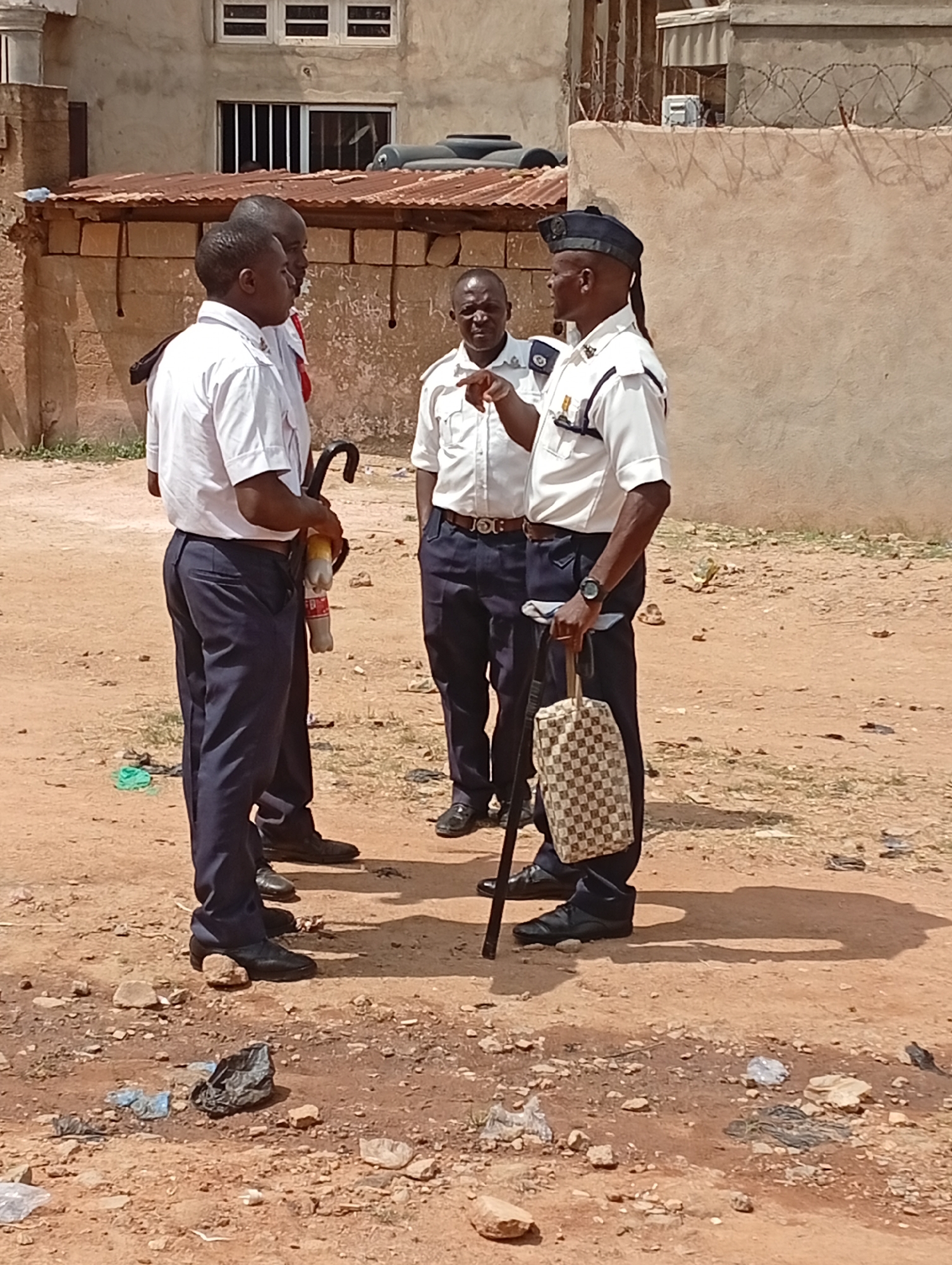 The image shows four uniformed men standing on a dirt road. The man on the right has a brimmed cap, a walking stick, and a checkered bag. He is pointing towards the man in the middle. The two men on the left have a fire extinguisher and a cane. The setting appears to be an urban area, given the nearby building. There is litter on the ground in the foreground.