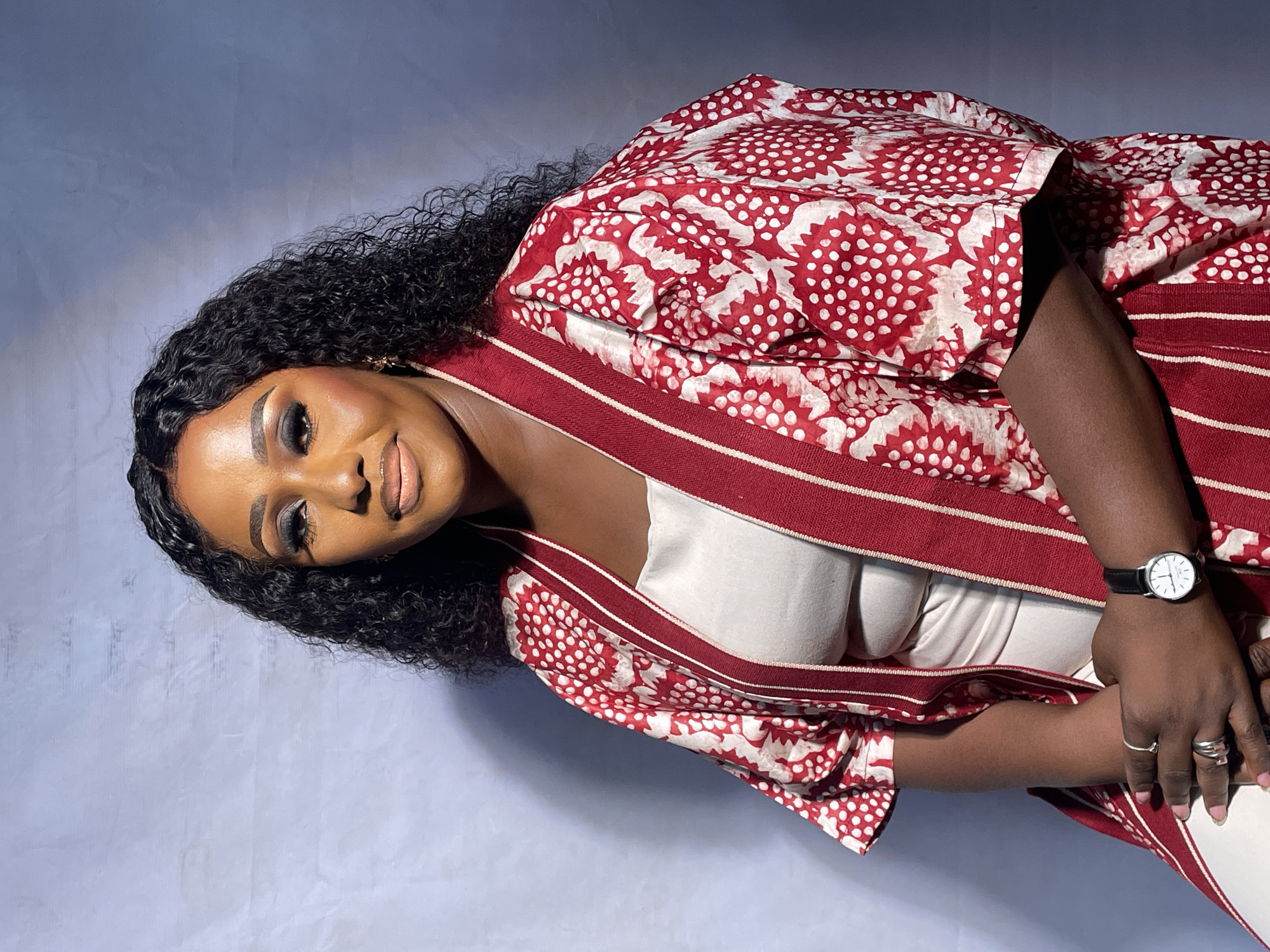 A dark-skinned woman with curly black hair is posing for a portrait in front of a plain light blue-grey background. She is wearing a white top and a red and white patterned robe with red striped trim. She has a wristwatch on her left wrist and rings on her fingers. Her makeup includes dark eye shadow and lipstick. It is not possible to verify the location of this scene in Jos, Nigeria based on visual details.