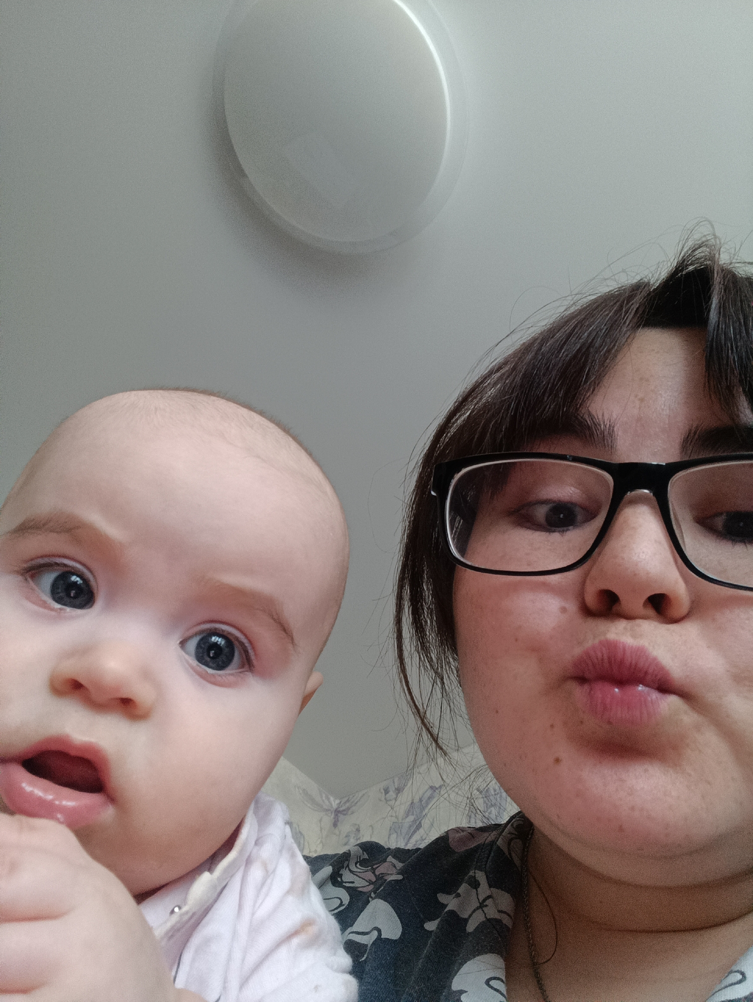 The scene is depicted indoors in the city of Noviy, Russia. The image shows a close-up, upward-angled view of two individuals: an adult female on the right and an infant on the left.
The adult female has dark hair and wears black-framed rectangular glasses. Her face is visible from the bridge of the nose downwards, displaying light freckling. Her lips are puckered in a reddish-pink color. She wears a dark-colored garment with a lighter pattern and a thin metal chain necklace.
The infant, positioned to the left, has light skin, prominent blue eyes, and sparse light hair. Its head, upper face, and upper chest are visible. The infant's mouth is slightly open, revealing the lower lip, and a small hand is held near its chin. The infant wears a light-colored top, possibly white or light pink, with a visible silver snap button.
The background consists of a light-colored ceiling, with a white, circular light fixture partially visible in the upper-center. Behind the lower portion of the individuals, a faint, light-colored patterned surface, possibly wallpaper or fabric, is present. The lighting is uniform.