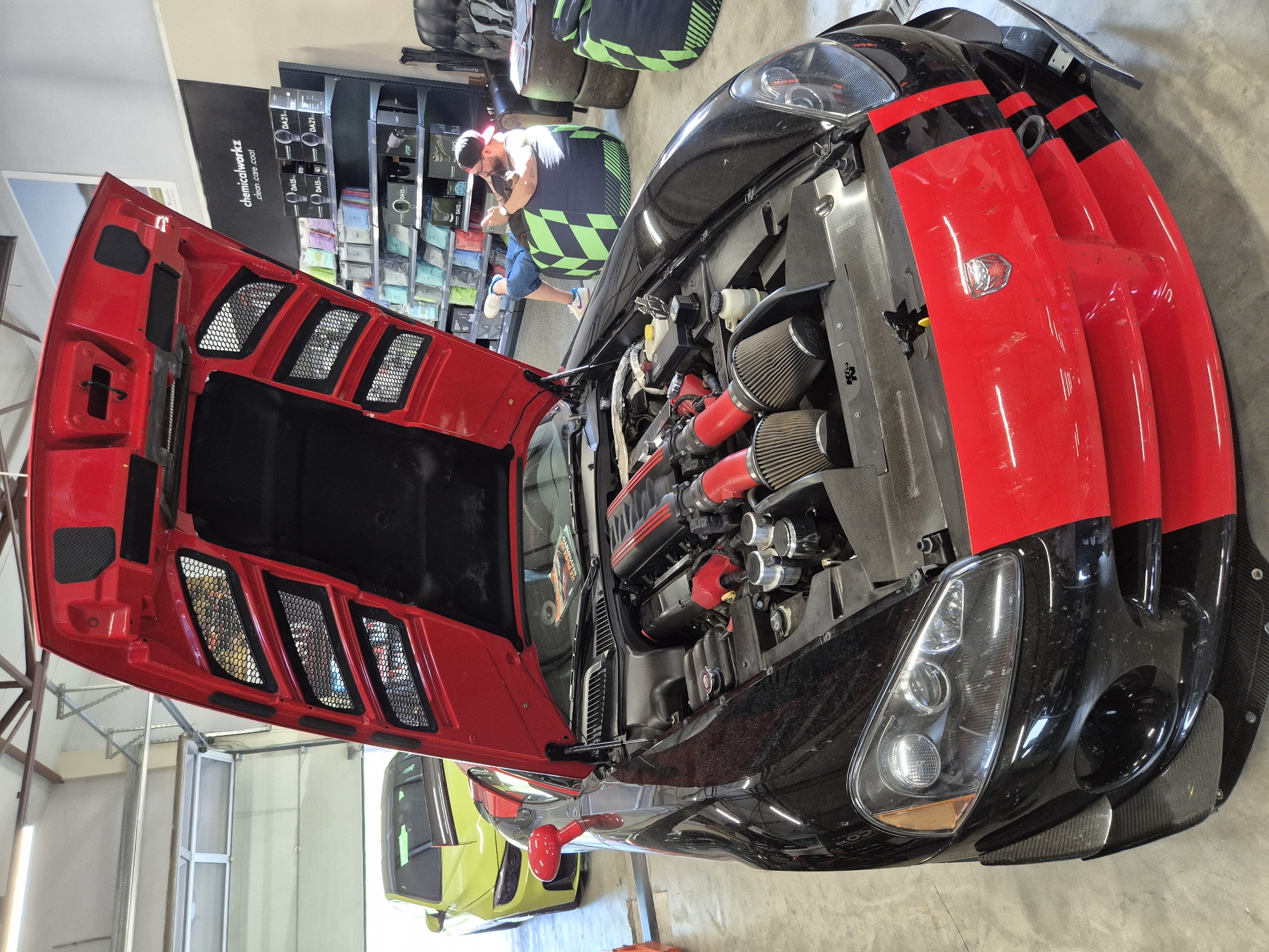 An indoor, eye-level photograph shows a black sports car with a prominent red stripe on its front, featuring its hood wide open, in what appears to be a garage or workshop located in Unknown, Unknown. The primary subject is a Dodge Viper with a black body and a red front fascia and hood stripe. The open hood reveals a modified engine bay with red intake tubing leading to dual K&N air filters. The car's front bumper displays carbon fiber aerodynamic elements, and a silver Viper emblem is centered on the red grille. In the left background, a bright yellow/lime green sports car is partially visible, featuring a black roof and a rear spoiler.
To the right of the main vehicle, an adult male with a beard, wearing a red baseball cap, dark t-shirt, blue shorts, and white sneakers, is seated on a large green and black geometrically patterned bean bag chair. He is holding a smartphone with both hands, appearing to be engaged with its screen. Behind him, a tall black shelving unit displays numerous car detailing products such as bottles and microfiber towels, branded with "chemicalworkz" and specific product codes like "DA15," "DA21," and "DA12." A large black banner above the shelves reads "chemicalworkz .clean .care .coat." Adjacent to the shelving unit is a black leather armchair and two stacks of tire-shaped seating, also covered in a green and black geometric pattern. The environment includes a light-colored concrete floor, white walls, and exposed metal ceiling trusses with fluorescent light fixtures. A large white roll-up garage door is partially open in the far background, allowing external light to enter.