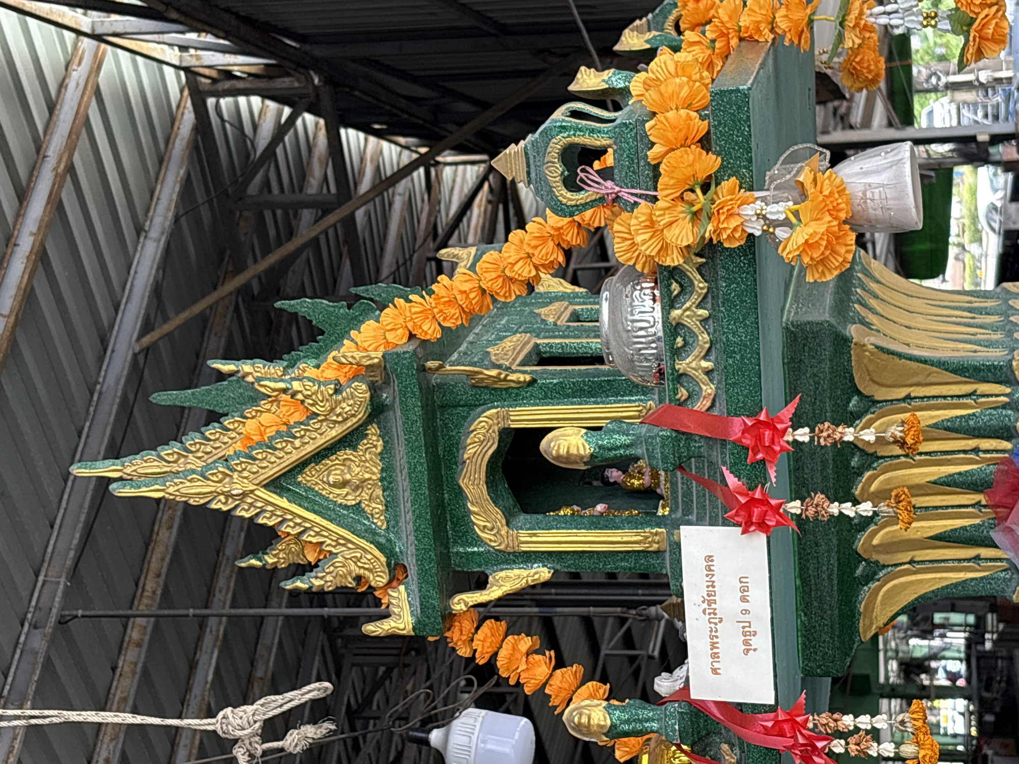 In Chalong, Thailand, a spirit house is adorned with orange flower garlands and red ribbons. The spirit house is predominantly green with intricate gold trim and decorative elements. Within an open archway, a small statue of a person, possibly a deity, is visible. A white sign with Thai script is hung below the archway. The structure appears to be under a metal roof, supported by a metal framework, suggesting it is located outdoors or in an open-air structure. A white light fixture hangs from the ceiling. Several strings of dried flowers with orange accents hang from the spirit house.