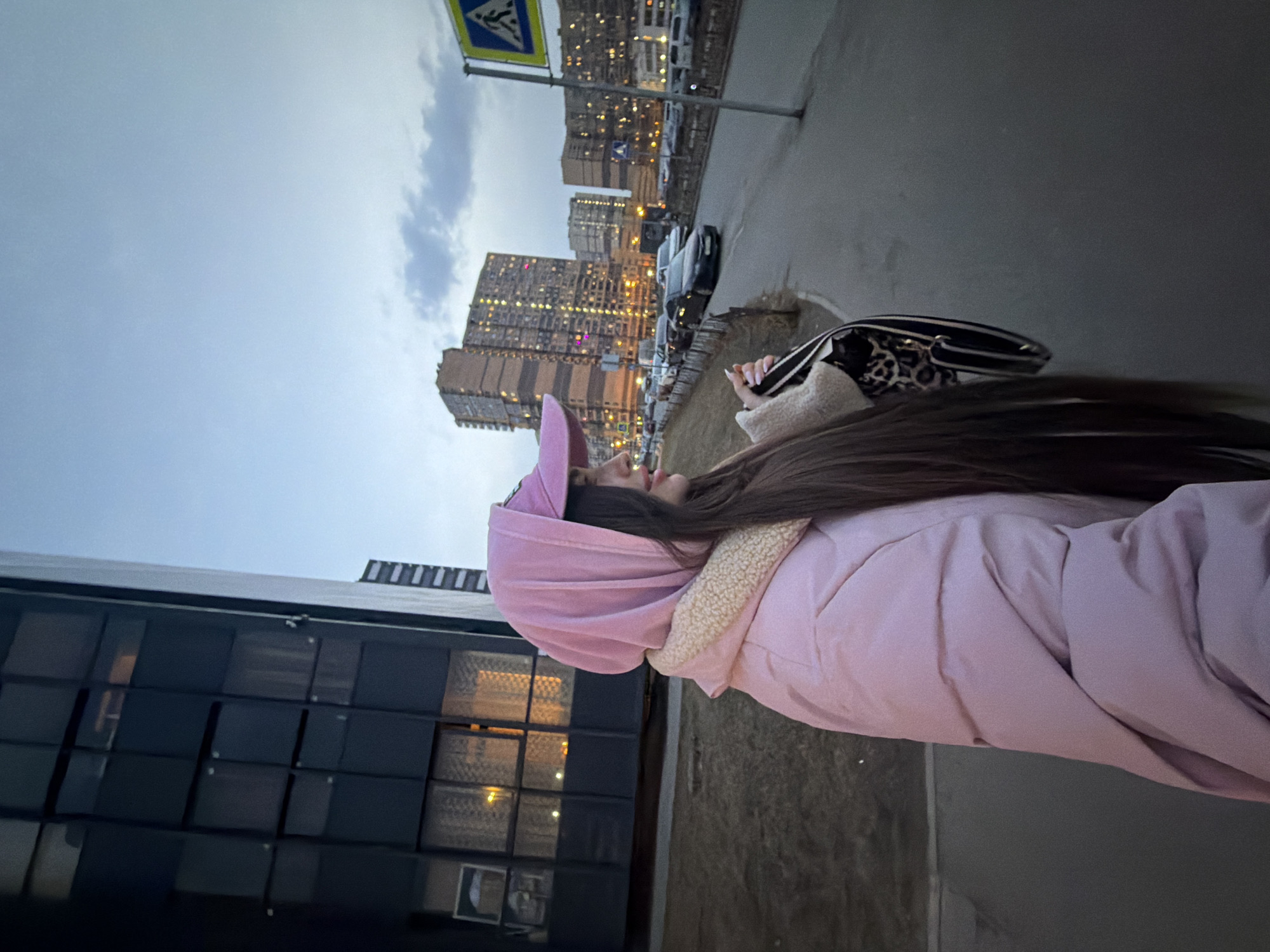 A person with long, dark hair wearing a pink hooded jacket stands on a sidewalk next to a multi-pane window, facing towards high-rise buildings. A crosswalk sign is visible, along with parked cars along a roadway, suggesting an urban setting. Based on architectural style and the crosswalk sign, the location is likely Saint Petersburg, Russia. The person is holding a black and white animal print purse. The sky is overcast.