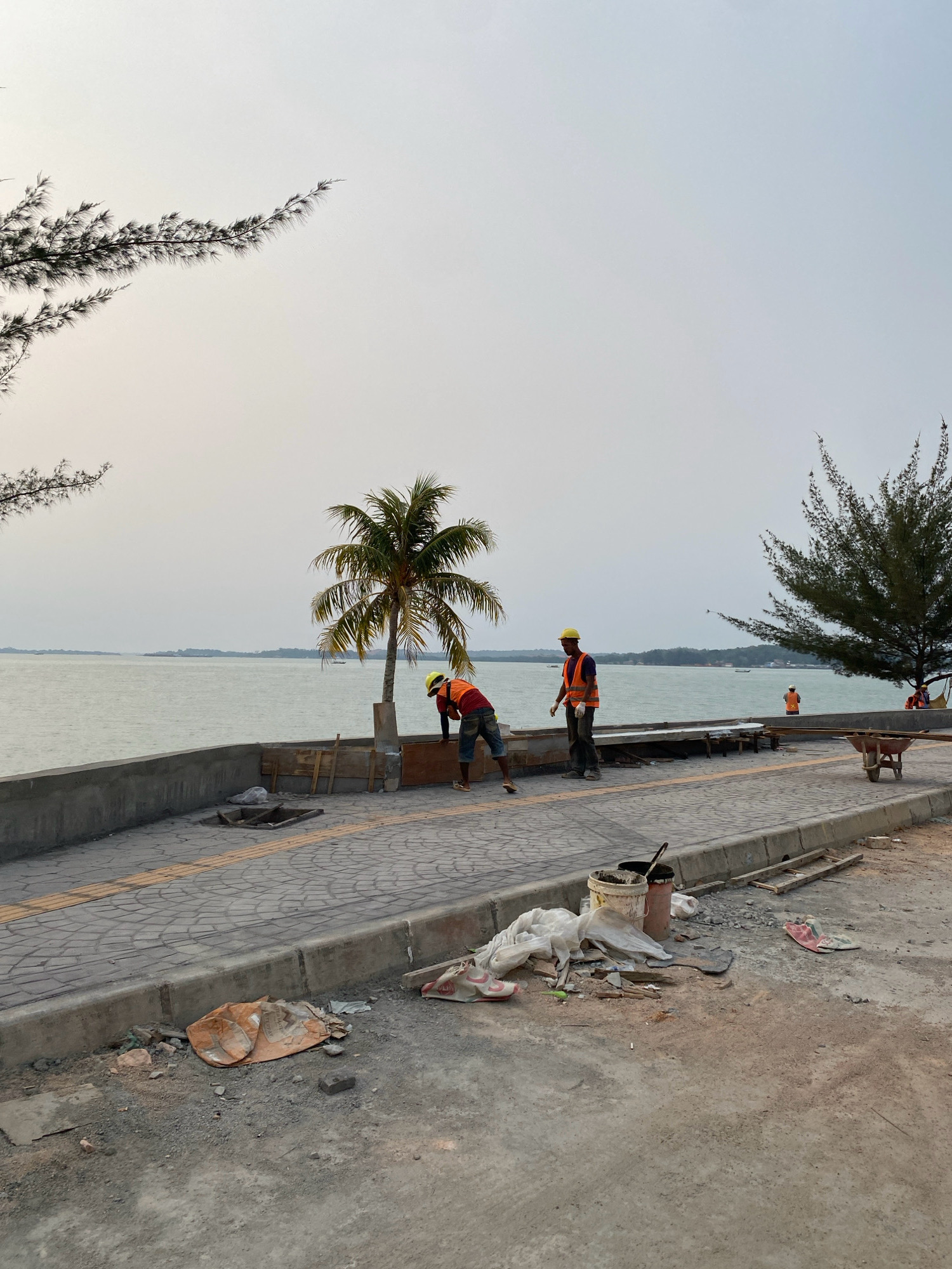 The image depicts a waterfront area undergoing construction in Tanjung Pinang, Indonesia. The scene is characterized by a paved walkway along a concrete sea wall, overlooking a calm body of water with distant landmasses. Two construction workers, dressed in orange safety vests and yellow hard hats, are visible. One is bent over, appearing to work on a wooden structure near a palm tree, while the other stands nearby, also engaged in construction-related activities. Debris, including plastic bags, buckets, and scattered materials, litters the unpaved ground in the foreground, suggesting ongoing work. A wheelbarrow is present further down the walkway, along with other construction supplies. Trees, including a large evergreen on the right and a palm tree near the workers, frame the waterfront. The sky is overcast, contributing to a muted, natural light across the scene. Several small boats are visible in the water in the distance. The overall impression is of a functional, albeit somewhat untidy, construction site in a coastal urban environment.