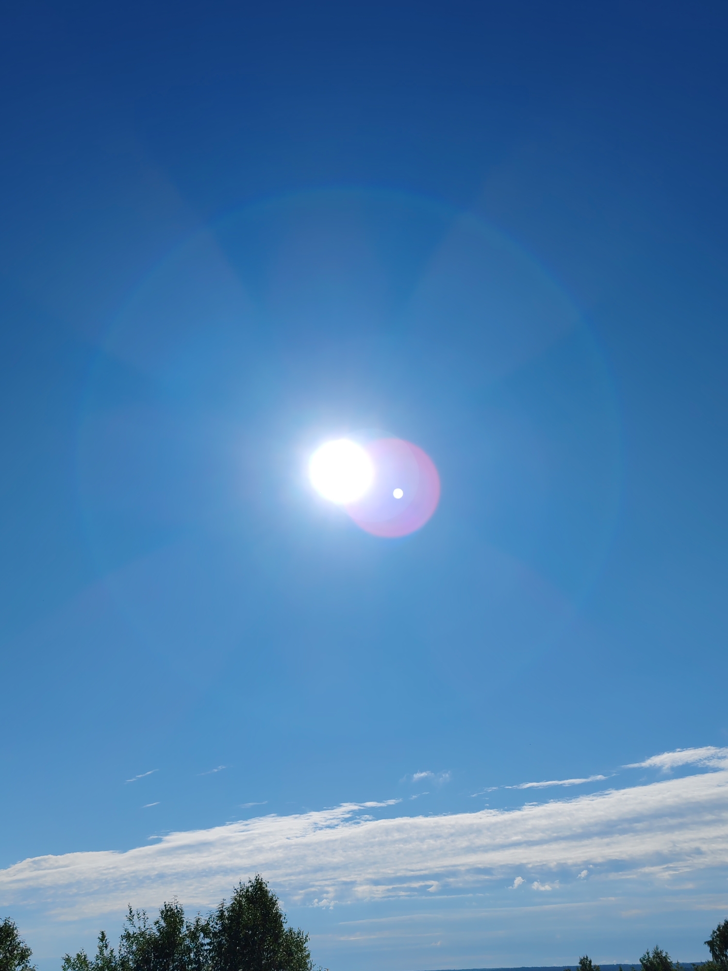 The image shows a bright sun with a lens flare, shining in a clear blue sky. There are some wispy clouds in the distance, and the tops of trees are visible along the bottom of the frame. The scene is likely outdoors in a rural or suburban area.