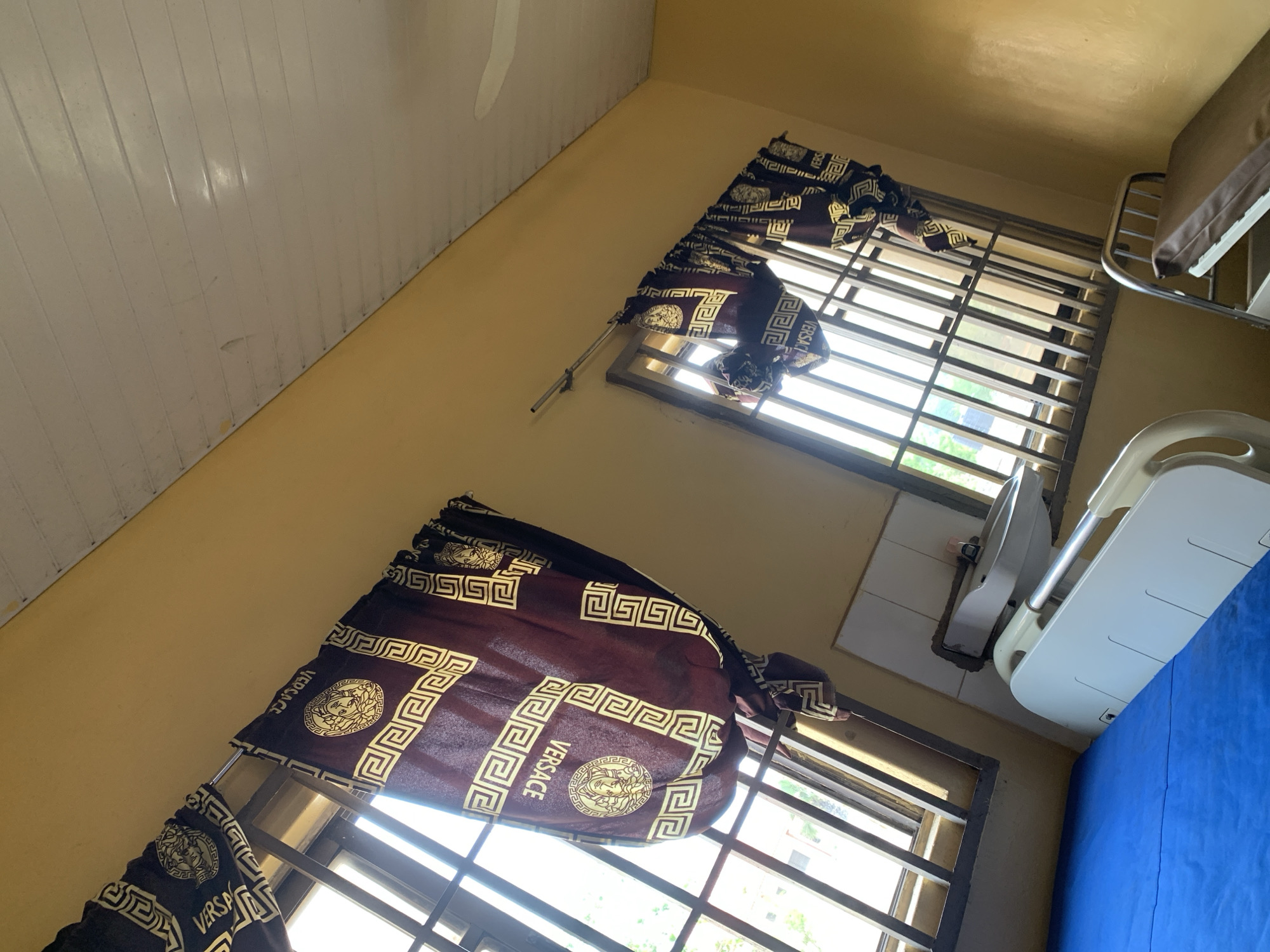 The image shows a room in Oko Erin, Nigeria. The room is painted yellow and has a white ceiling. There are two windows, each covered with brown curtains that have a Versace pattern. The windows have metal bars. A hospital bed with blue sheets is partially visible on the right side of the frame. Next to the bed is a sink. The scene appears to be a hospital room.