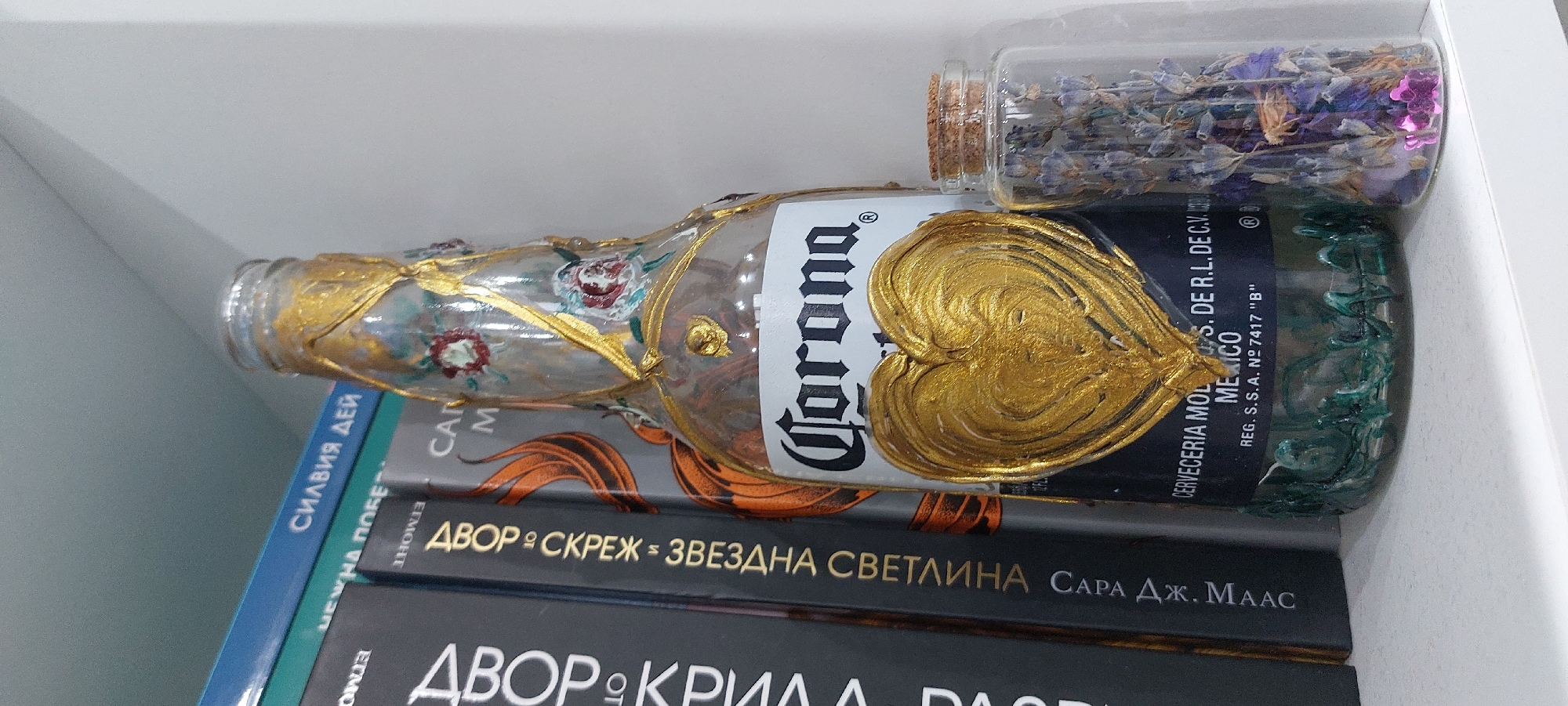 The image displays a creatively decorated Corona beer bottle resting horizontally on a stack of books, positioned on a white shelf in Yagodovo, Bulgaria. The clear glass bottle is adorned with elaborate gold paint, forming a prominent heart shape and delicate floral patterns with red roses. The bottom portion of the bottle is covered in a textured, dark green coating.
Adjacent to the decorated bottle, a small clear glass vial is visible, filled with dried purple and blue flowers, likely lavender, mixed with some pink glitter. Both the bottle and the vial appear to be stationary decorative items.
Beneath these objects, several books are stacked. The most visible book, dark with gold lettering, reads "ДВОР ⁕ СКРЕЖ ⁕ ЗВЕЗДНА СВЕТЛИНА САРА ДЖ. МААС" (Court of Frost and Starlight by Sarah J. Maas). Below it, another dark book spine shows "ДВОР ⁕ КРИЛА ДА" (Court of Wings and Ruin). A partially visible blue book spine reveals "СИЛВИЯ ДЕЙ" (Silvia Day). Other partial texts from the books include "ETMOНТ", "NEWHA ПОБЕ", "CAN", and "M".
The Corona bottle's label clearly shows "Corona ®", "CERVECERIA MOLS. DE R.L. DE C.V.", "MEXICO", and "REG. S.S. A. Nº 7417 "Β"". The scene is an indoor setting, with no activity taking place, and no specific time of day or weather conditions are discernible.