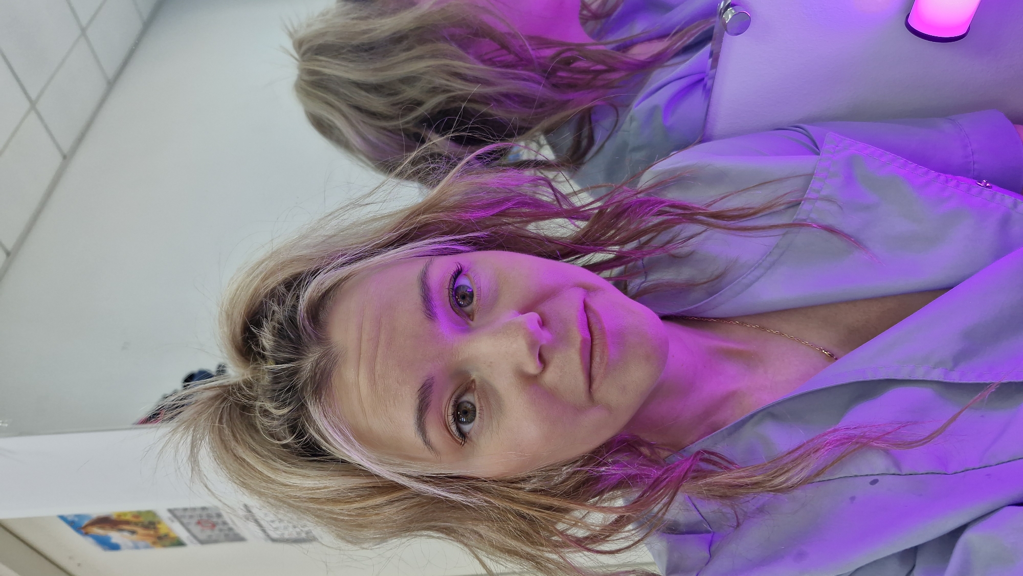A woman with blonde hair and green eyes is looking directly at the camera. She is wearing a light blue button-down shirt and a gold necklace. Her reflection is visible in the mirror behind her. The scene is illuminated by purple lighting. A white wall with tile-like patterns and some other blurry objects are also visible in the mirror. The location is likely indoors, given the presence of the mirror and tiled wall. However, the exact location in Otchor, Russia, cannot be determined from this image alone.