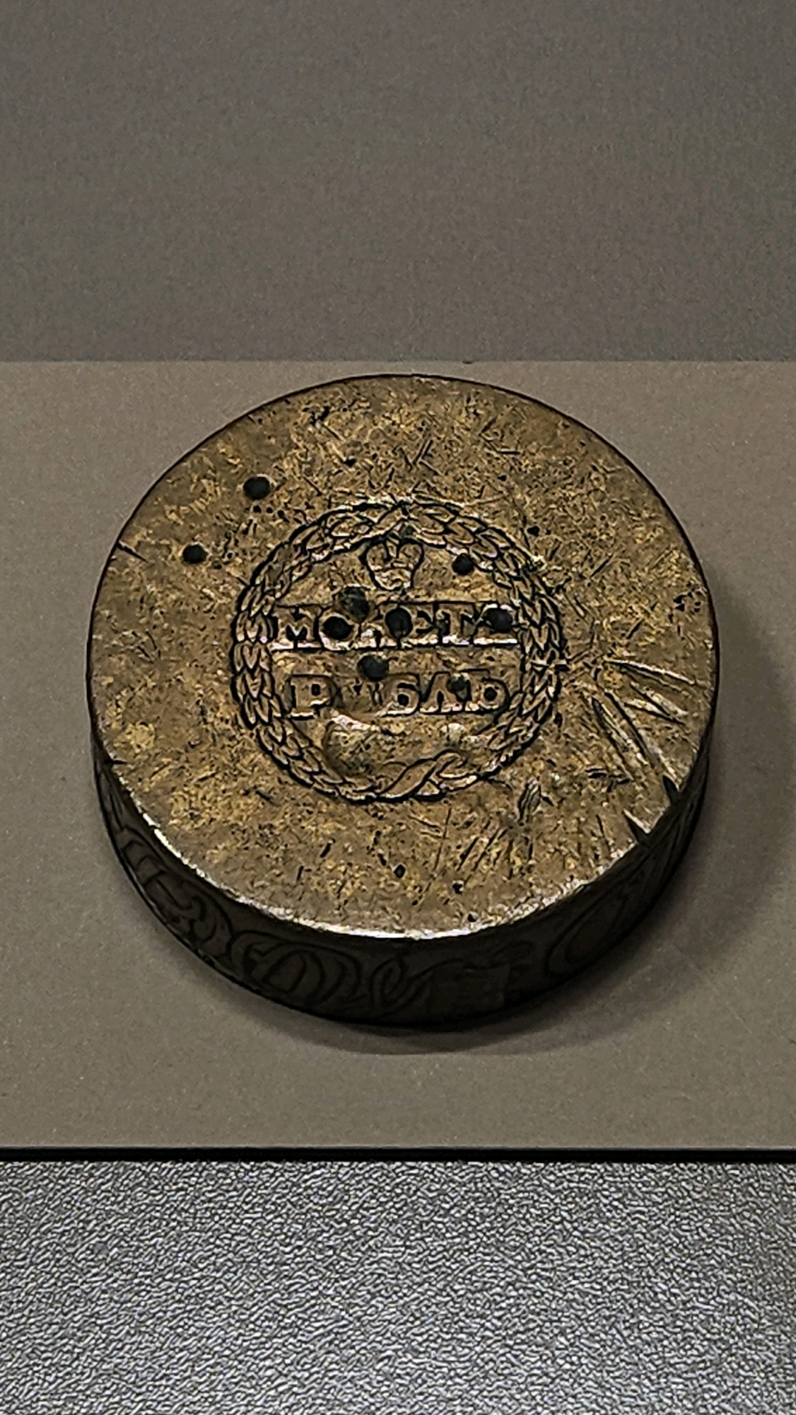 The image shows a close-up, eye-level, slightly angled shot of a weathered, circular, brass object, likely a coin or medal, resting on a neutral-colored surface. The object's face is heavily textured with scratches and small indentations, giving it an aged appearance. In the center of the object, a circular motif is visible, featuring what appears to be a crown at the top, enclosed by a laurel wreath. Below the crown, the inscription "МОНЕТА" (Moneta) is clearly legible, followed by "РУБЛЬ" (Rubl) in a similar font. The side of the object has a decorative, engraved pattern. The background is a muted grey surface that extends upwards, with a textured dark grey surface below. There are no people present in the image. The scene does not provide any specific information that would allow for the localization of the object to the city of Ulyanovsk, Russia.