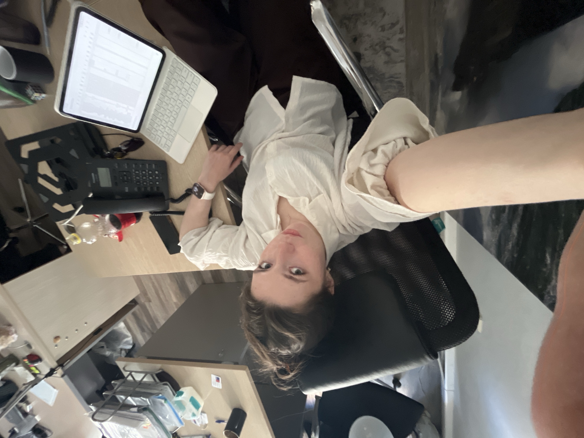 A woman sits in a black office chair at a desk, on which sits an Apple iPad, a phone, and other assorted office supplies. The background is partially obscured, but there is a white wall with a painting of a landscape. The location cannot be determined.