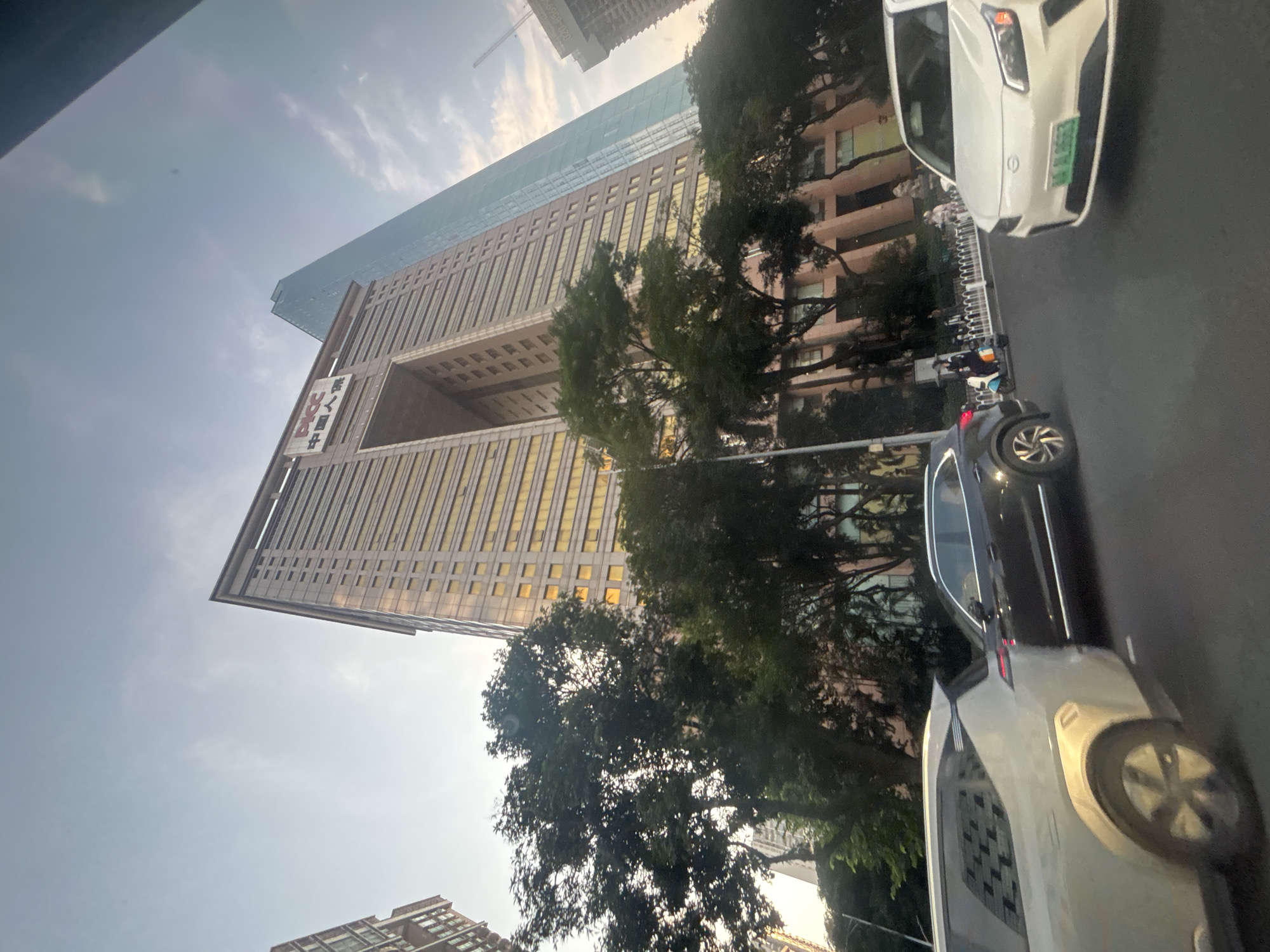 This is a wide-angle, eye-level shot taken from a vehicle traveling on a city street in Tianhe District, China. The image captures a mix of modern architecture and urban street life.
Dominating the background is a very tall, contemporary skyscraper with a distinctive large archway near its peak. The building has numerous floors with golden-tinted windows, reflecting the sky. To the right of this skyscraper, another much taller, sleeker glass skyscraper tapers to a point, also reflecting the sky. The name "PICC" is visible in red lettering on the side of the archway building, along with Chinese characters.
In the middle ground, lush green trees partially obscure the lower floors of the buildings. The street level shows a white electric vehicle in the foreground on the right, its license plate visible. To its left, a dark SUV is also on the road, and further left, the rear of a light-colored SUV is visible. Several pedestrians are present on the sidewalk behind a white fence, with some on scooters. The sky is a pale blue with some scattered clouds. The overall lighting suggests it is late afternoon or early evening.