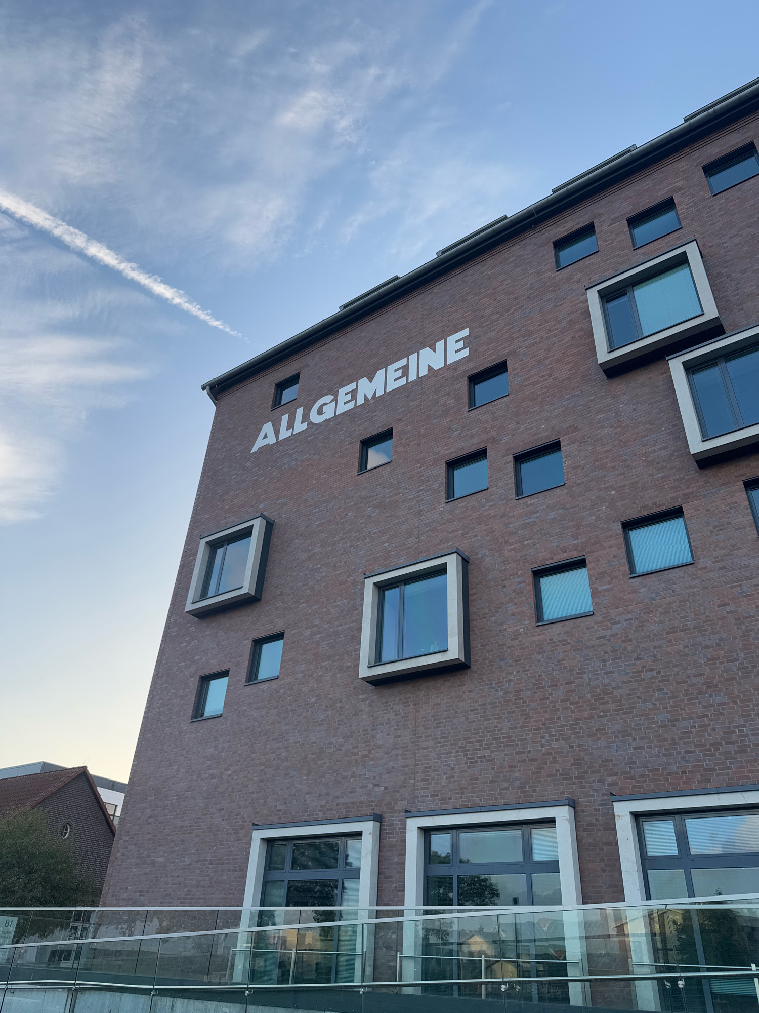 The image shows a modern, brick-faced building in Cleves, Germany, with the word "ALLGEMEINE" in large white letters across its facade. The building has a distinctive architectural style with numerous square and rectangular windows of varying sizes, some protruding outwards. The sky above is blue with a few wispy clouds and a contrail from an airplane. Below the building, a contemporary glass and metal railing separates the structure from a lower level. The overall impression is one of modern urban architecture against a daytime sky.