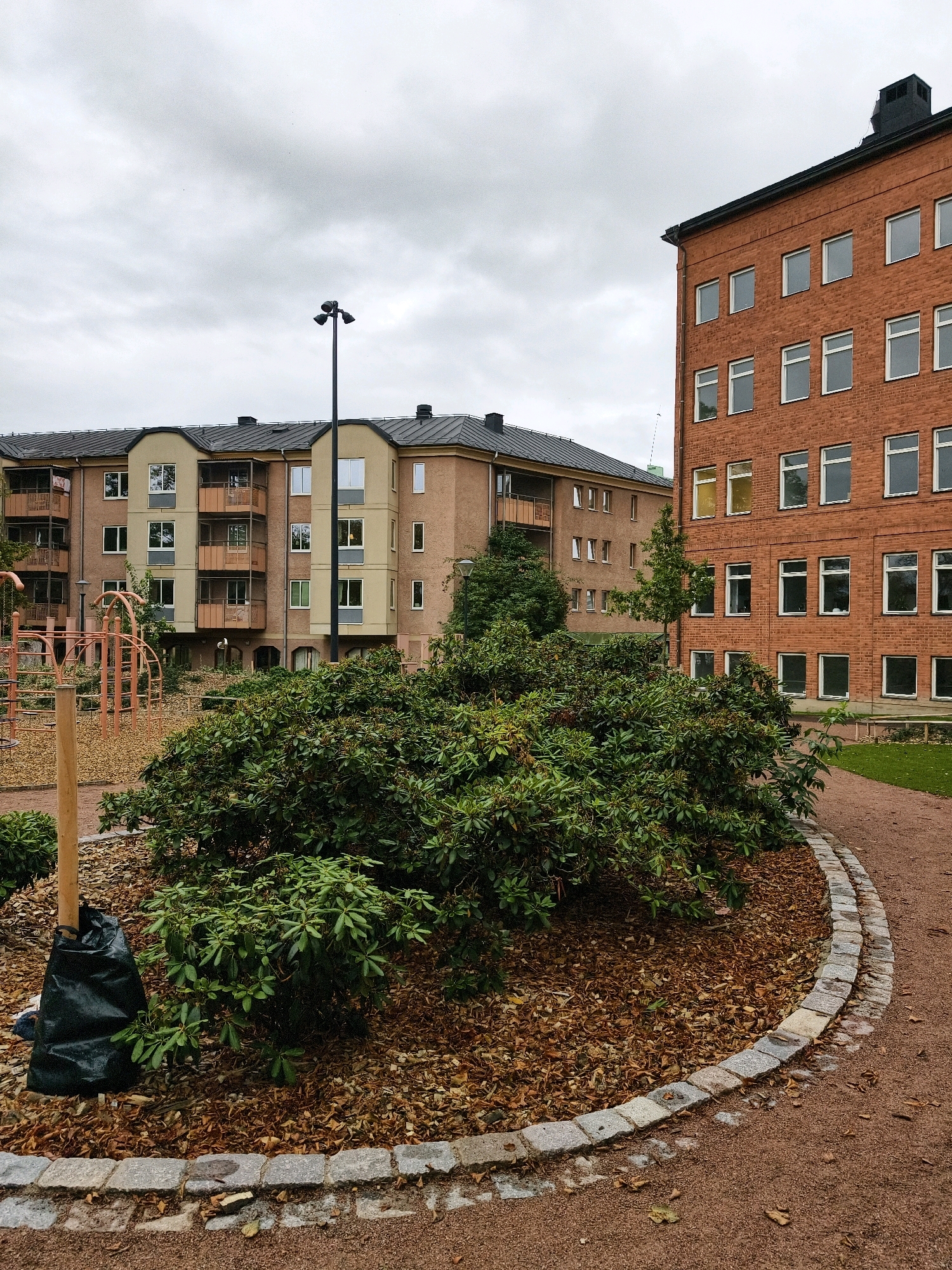 The image shows a view of an apartment complex and a red brick building in Linköping, Sweden. In the foreground, there is a circular garden bed with a large, dense bush. A wooden stake wrapped in black plastic is visible to the left of the bush. The garden bed is surrounded by a curved stone border and covered in mulch and fallen leaves. A gravel path curves around the garden bed and leads to the right.
In the middle ground, two apartment buildings are visible. The building on the left is multi-story with a light-colored facade and dark gray roofs. It features balconies with glass enclosures. The building on the right is made of red brick and has multiple floors with many windows. A tall black lamp post with three lights is located between the two buildings.
In the background, more of the apartment complex and a tree are visible. The sky is overcast and cloudy. The scene appears to be an outdoor common area or courtyard within a residential development.