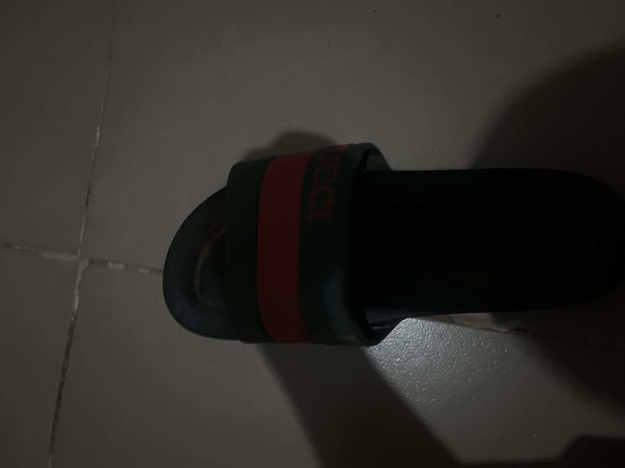 A black Gucci slide sandal is pictured on a tiled floor. The sandal has a dark green strap with a red stripe across the top, and the Gucci logo is visible in red letters on the strap. The background is a dimly lit, tiled surface, suggesting an indoor setting. The image is taken from a high angle, with the sandal positioned in the center of the frame. The overall lighting is poor, casting shadows on the floor and wall. The location is specified as Jalingo, Nigeria, though no specific landmarks or context are provided to visually confirm this.
