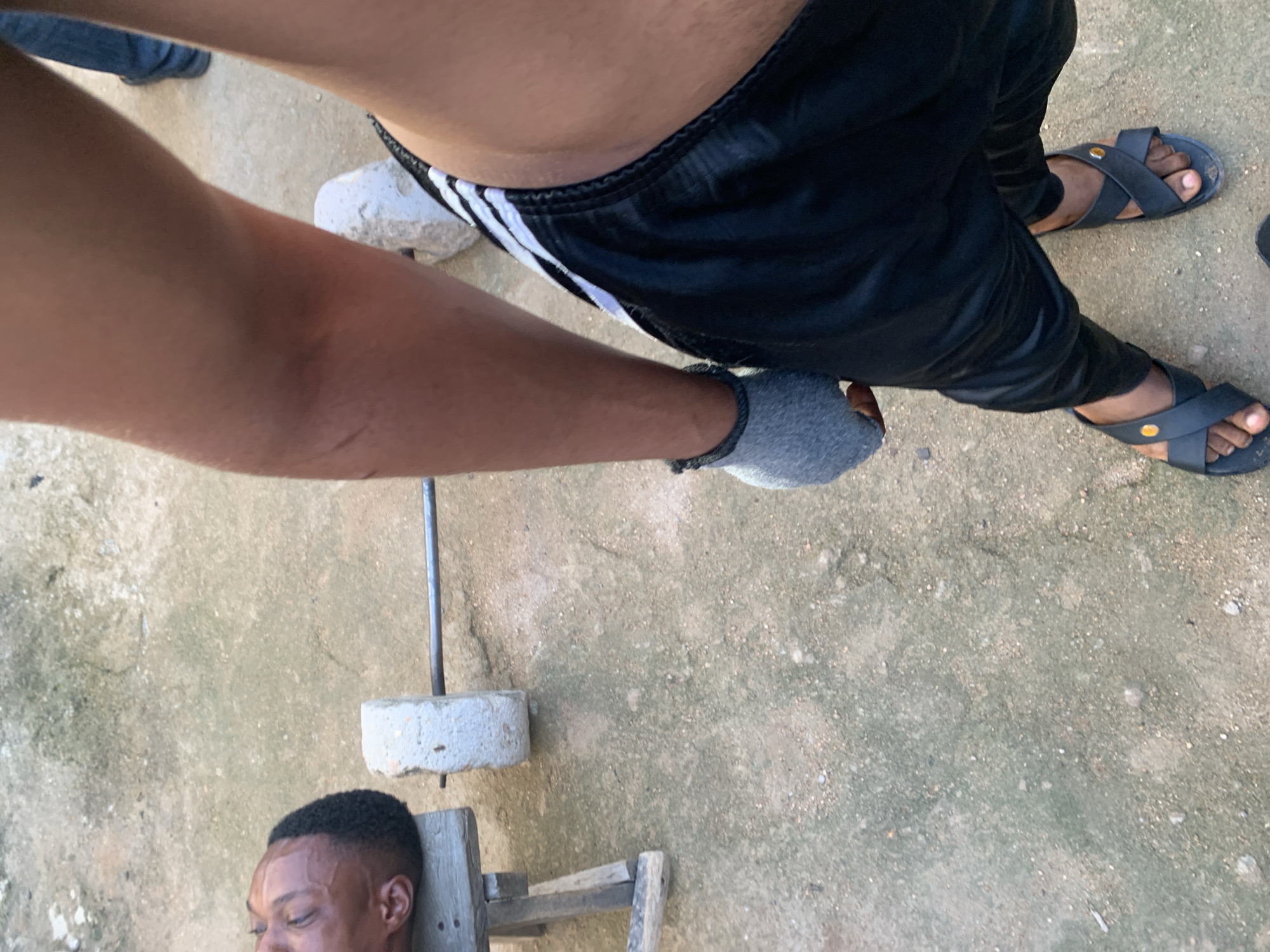 In Jauro Yinu, Nigeria, an individual is captured from the waist down, wearing black athletic shorts with white stripes and dark sandals. They are holding a weightlifting bar with concrete weights attached, suggesting a workout session. The scene is set outdoors on a gravelly ground. To the left, a person's head rests on a wooden support, their eyes closed, possibly asleep or resting.