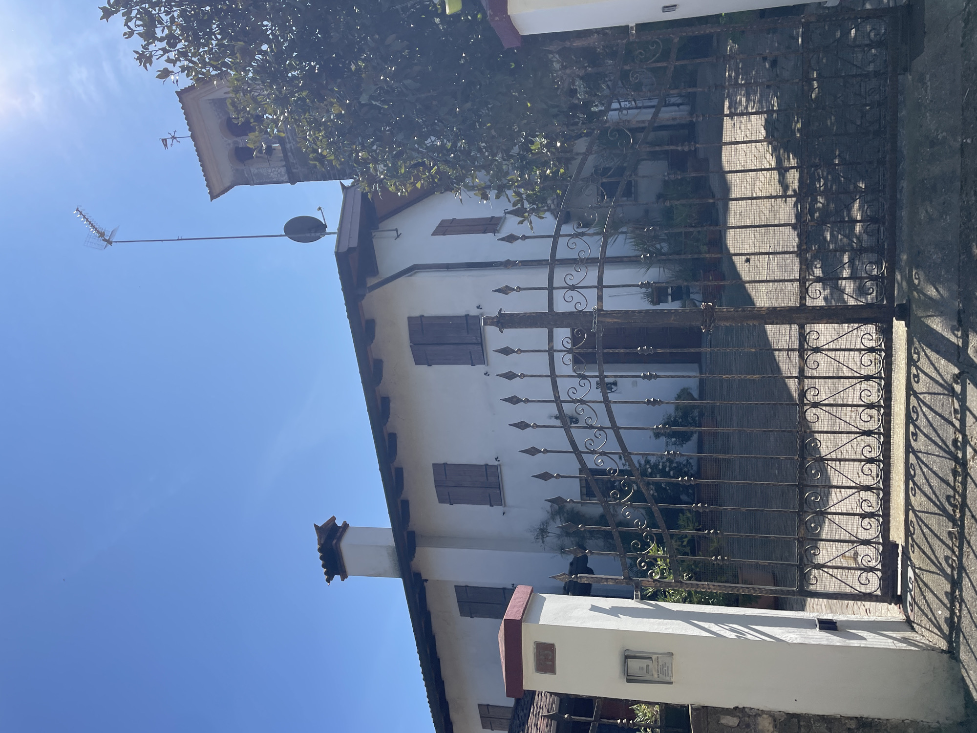 A low-angle shot shows a white, multi-story building with brown shutters behind a wrought-iron gate in San Lorenzo, Italy. The building has a satellite dish and antenna on its roof. A church tower is visible behind the building. The sky is clear and blue. The gate obscures much of the view of the building and the courtyard behind it. Sunlight is visible.