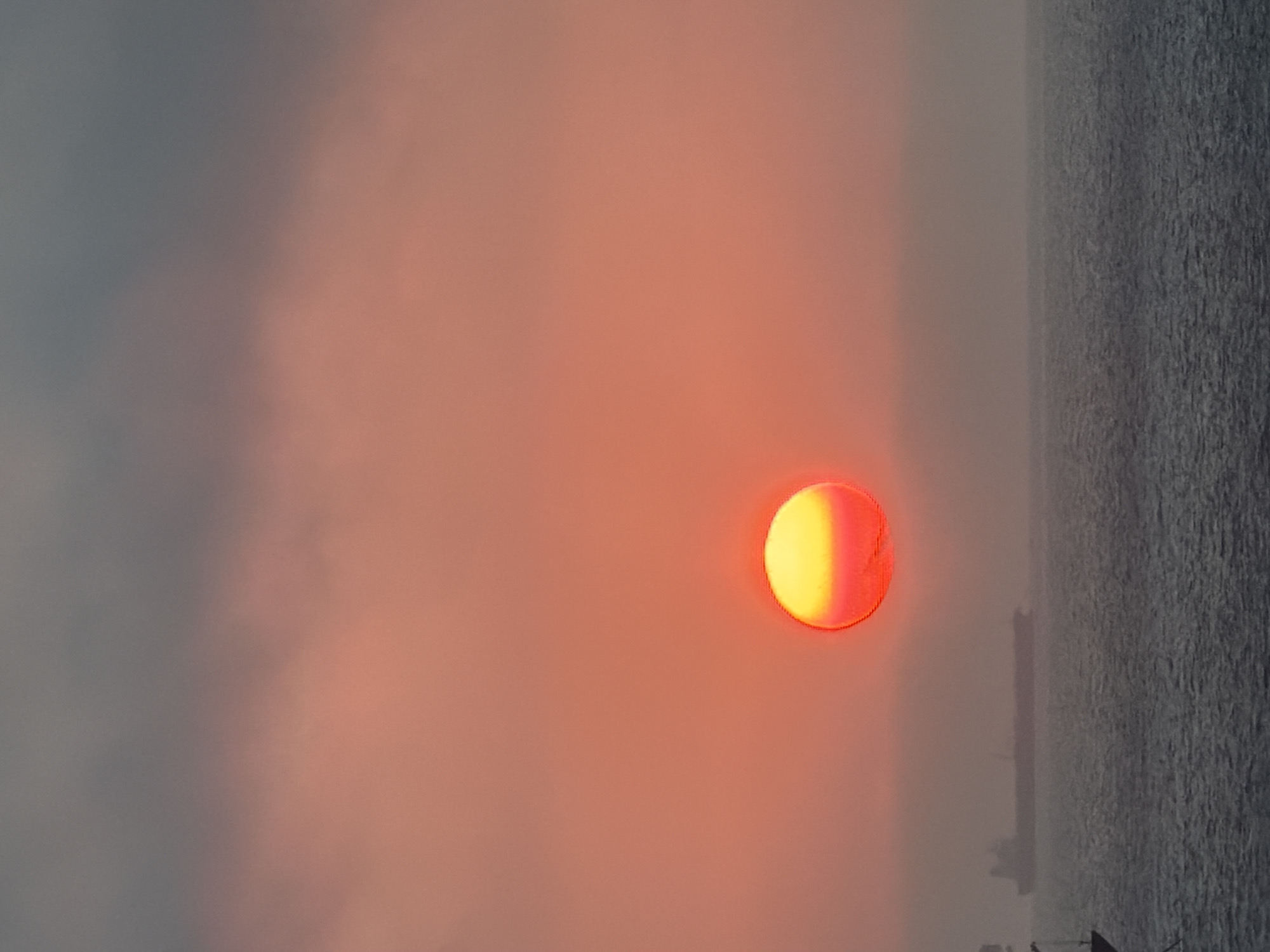 The image shows a sunset over water. A round, glowing sun, partially obscured, is visible near the center. The sky is a blend of orange and grey hues, transitioning to a darker grey water body on the right side of the image. There appears to be a ship in the distance but the image is unfocused. The location, city, and specific details regarding people, interactions and actions are unknown.