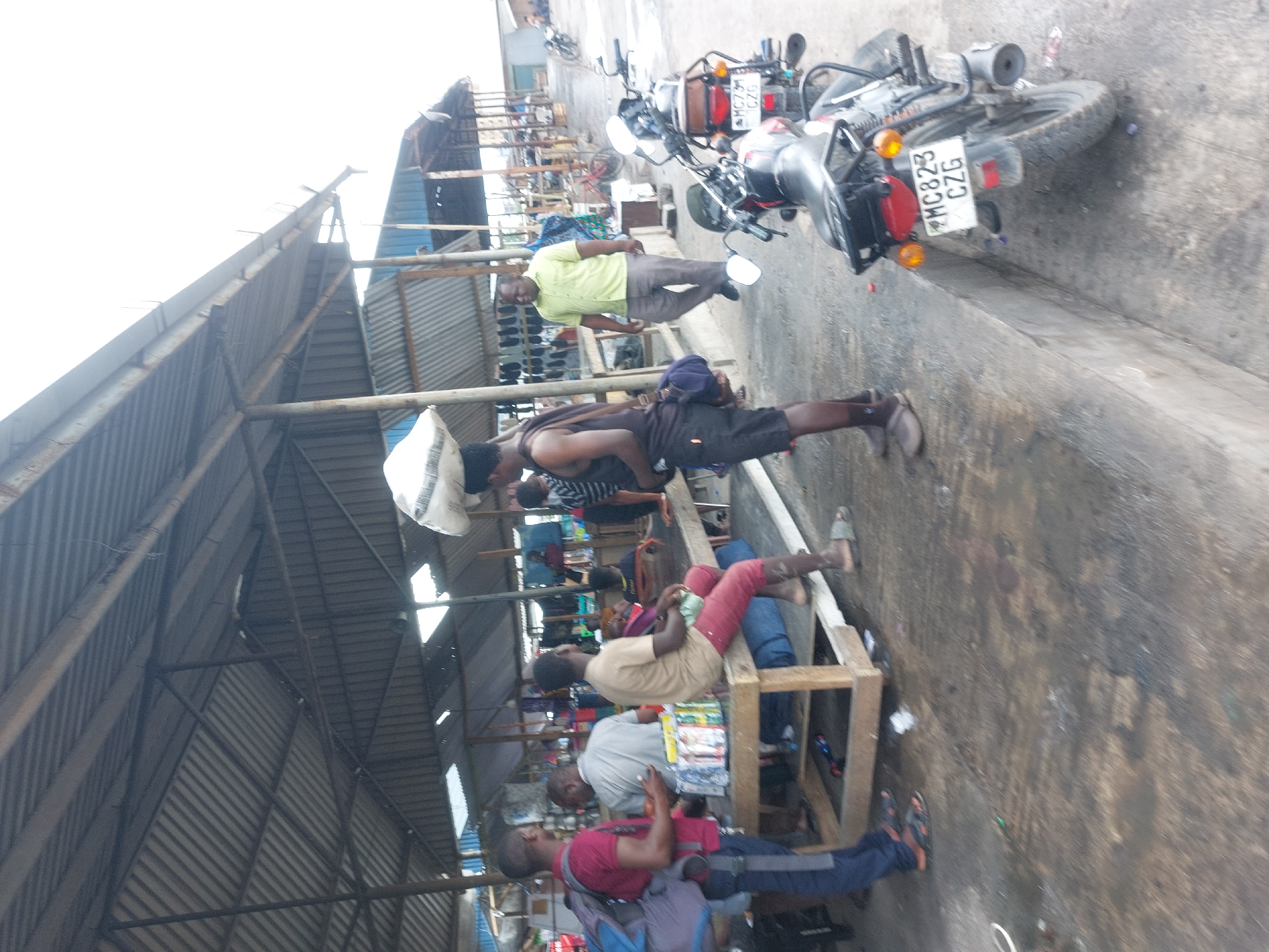 This image captures a busy outdoor scene, likely a market or transport hub, in Dar es-Salaam, Tanzania, under a series of corrugated metal shelters. The environment is utilitarian, characterized by metal-framed structures with grey, corrugated roofs and walls. The ground is a worn, dark-colored concrete or asphalt.
Several people, predominantly men, are visible. In the mid-ground, a man in a lime green shirt and grey pants stands, looking towards the right. To his left, another man in a dark tank top and shorts is seated on a low wooden platform, balancing a large white sack on his head. Further into the foreground (bottom half of the image when viewed upright), more men are seated on wooden benches or platforms. One man wears a light brown shirt and reddish-brown pants, while another in a grey shirt sits facing the viewer, with a stack of magazines or books in front of him. A third man, dressed in a red shirt and dark blue pants, is seated lower, possibly on the ground, with a backpack beside him.
Two motorcycles are parked on the right side of the scene. One is black with chrome accents, displaying a visible license plate: "MC823 CZG." The overall activity suggests a casual gathering or waiting area, typical of a bustling market environment. The lighting is bright, indicating daytime, but the sky is overexposed, making the exact weather conditions unclear. No strong emotions are discernible from the individuals.