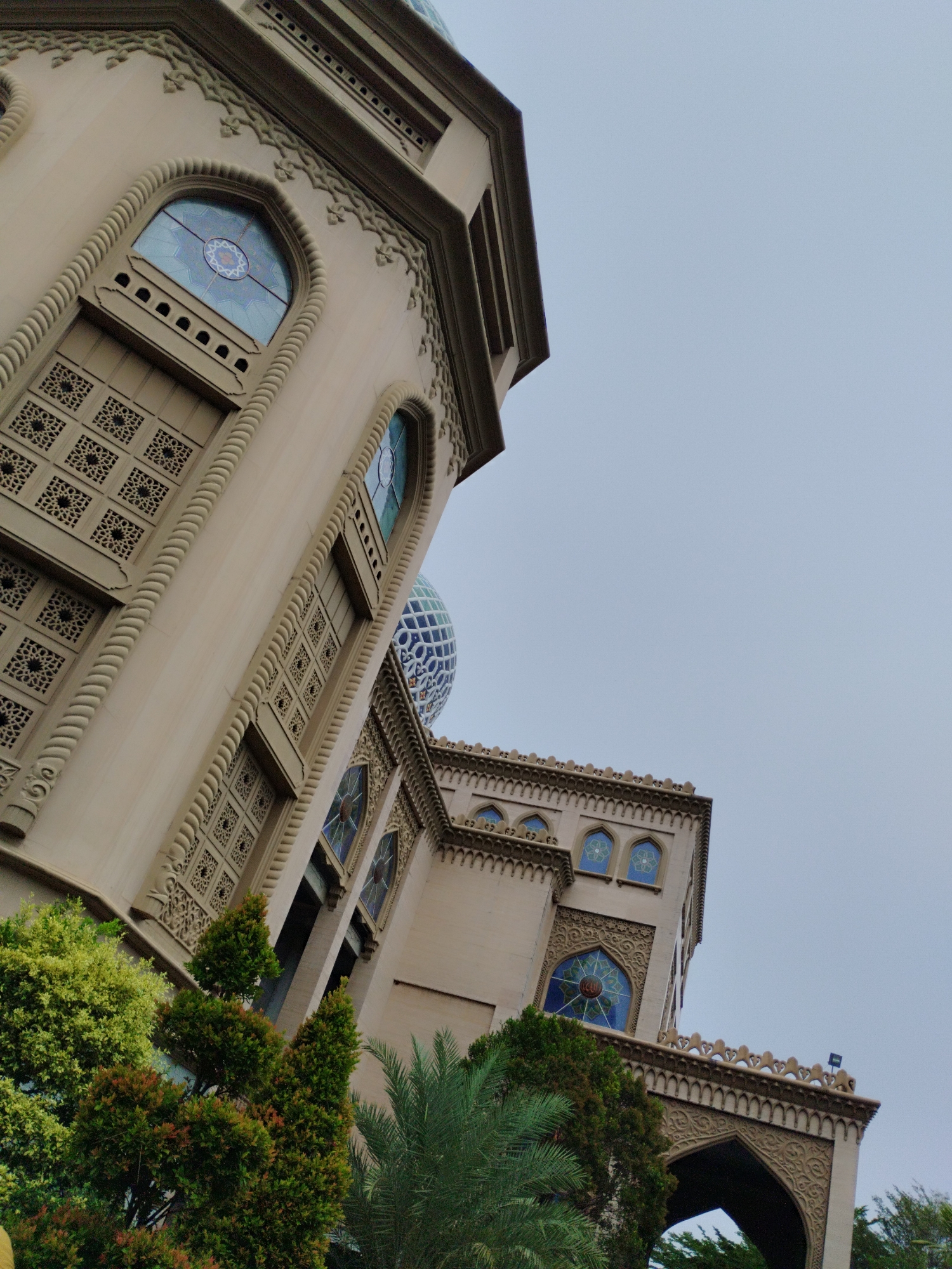 The image shows a low-angle view of a mosque with distinct Islamic architecture. The facade is light beige and features intricate decorative carvings, arched windows with stained glass, and patterned screens. Two prominent structures are visible: a taller tower on the left and a slightly shorter, wider building on the right, connected by an arched walkway. A large, patterned blue dome is visible behind the right-hand building. Lush green trees and foliage are in the foreground, partially obscuring the lower sections of the mosque. The sky is overcast and pale blue. The mosque is identifiable as the Baiturrahim Mosque in Lhokseumawe, Aceh, Indonesia.