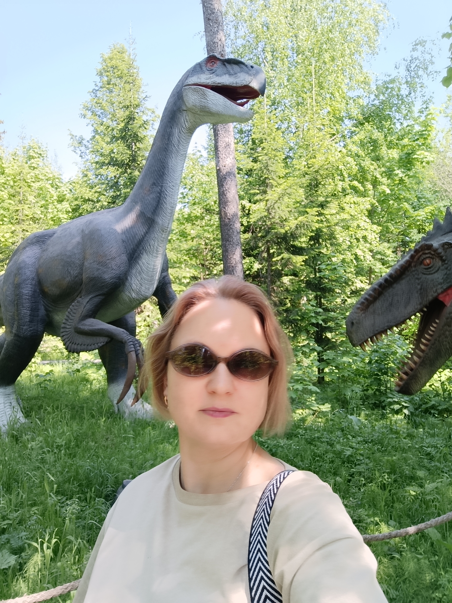 A woman wearing sunglasses is standing in front of two dinosaur statues in a park. The woman is wearing a light-colored shirt and a purse with a black and white strap. The statue behind her on the left is gray and standing on two legs, and has a long neck and large claws. The statue behind her on the right is green and has a large mouth with teeth. The park is grassy with many trees. The location is Izhevsk, Russia.