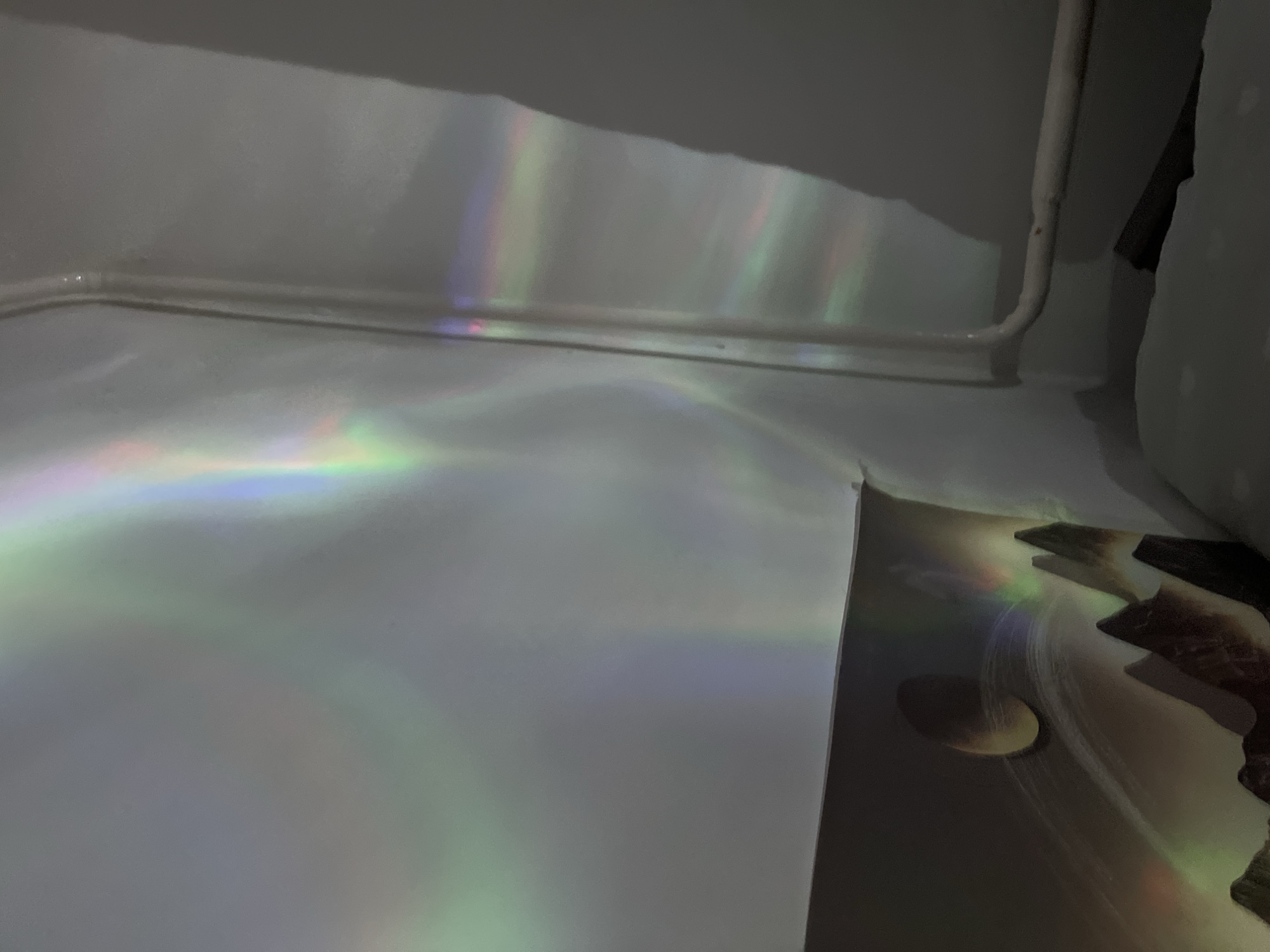 This image depicts an indoor scene, likely a room, with light effects projected onto the walls and ceiling. The light appears to be creating a spectrum of colors, resembling an aurora borealis or rainbow effect, with bands of green, blue, pink, and yellow visible. The walls are a neutral color, likely white or off-white, and there is a pipe or conduit running along the right wall. In the lower left portion of the image, a dark surface is visible, possibly a poster or a wall decoration, featuring a silhouette of mountains and a circular object, perhaps a planet or moon. The overall impression is one of ambient lighting and possibly a decorative light fixture. There are no people or animals present in the image. The location is not specified in the image itself.