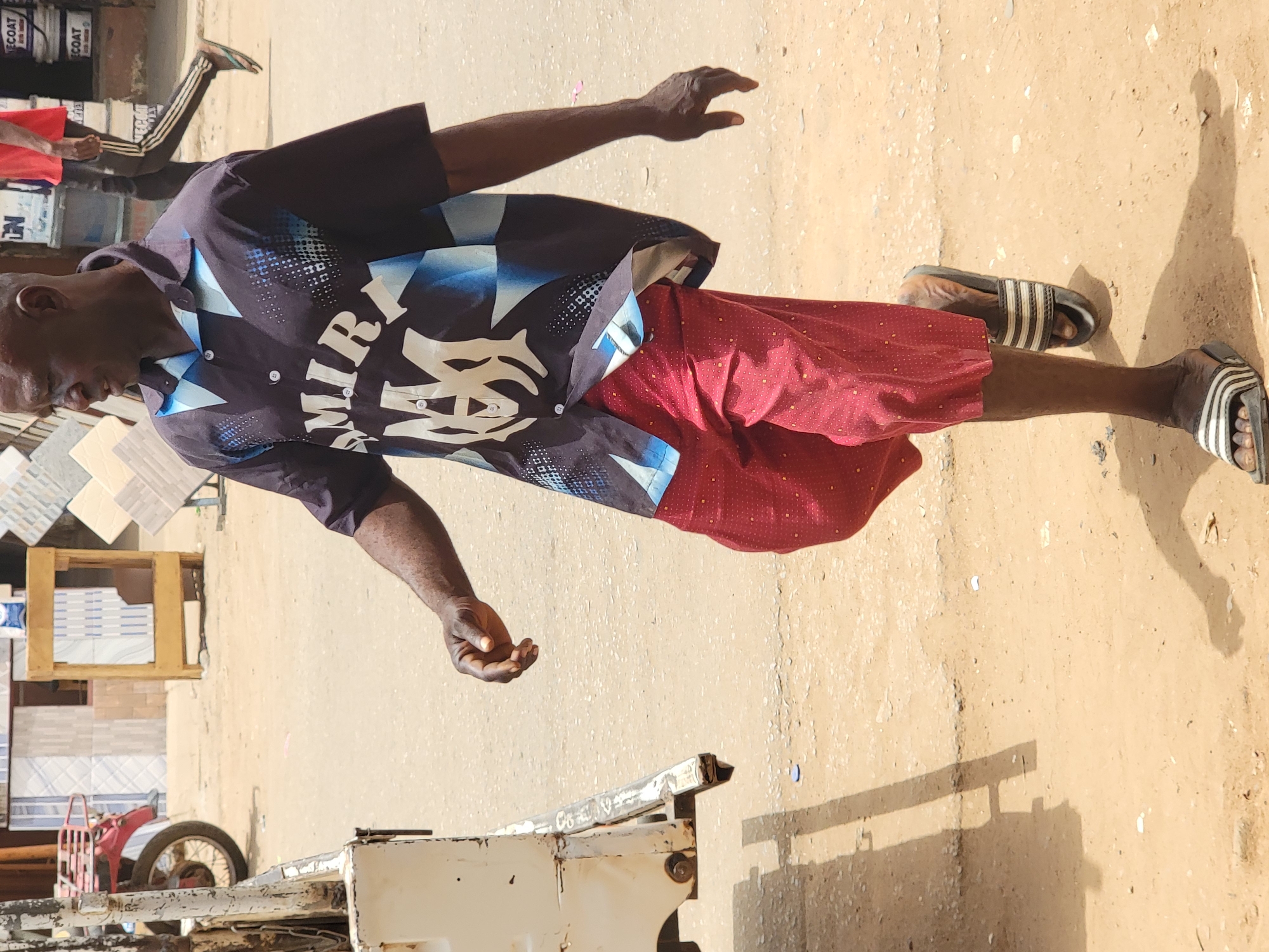 An adult male stands on a pale brown, cracked, and uneven pavement, facing slightly away from the viewer. He wears a dark, short-sleeved button-down shirt with blue and white geometric designs and the word "WIRI" in white across the chest. He also wears red shorts with a small dot pattern. He is wearing open-toed sandals with black and white stripes. His arms are outstretched with his hands open. In the background, there is a partially visible cart, various building materials and what looks to be an assortment of tiles and paint. Another person is partially visible in the top left corner of the image. The ground is light, suggesting a sunny outdoor environment. The location is Maiburiji, Nigeria.