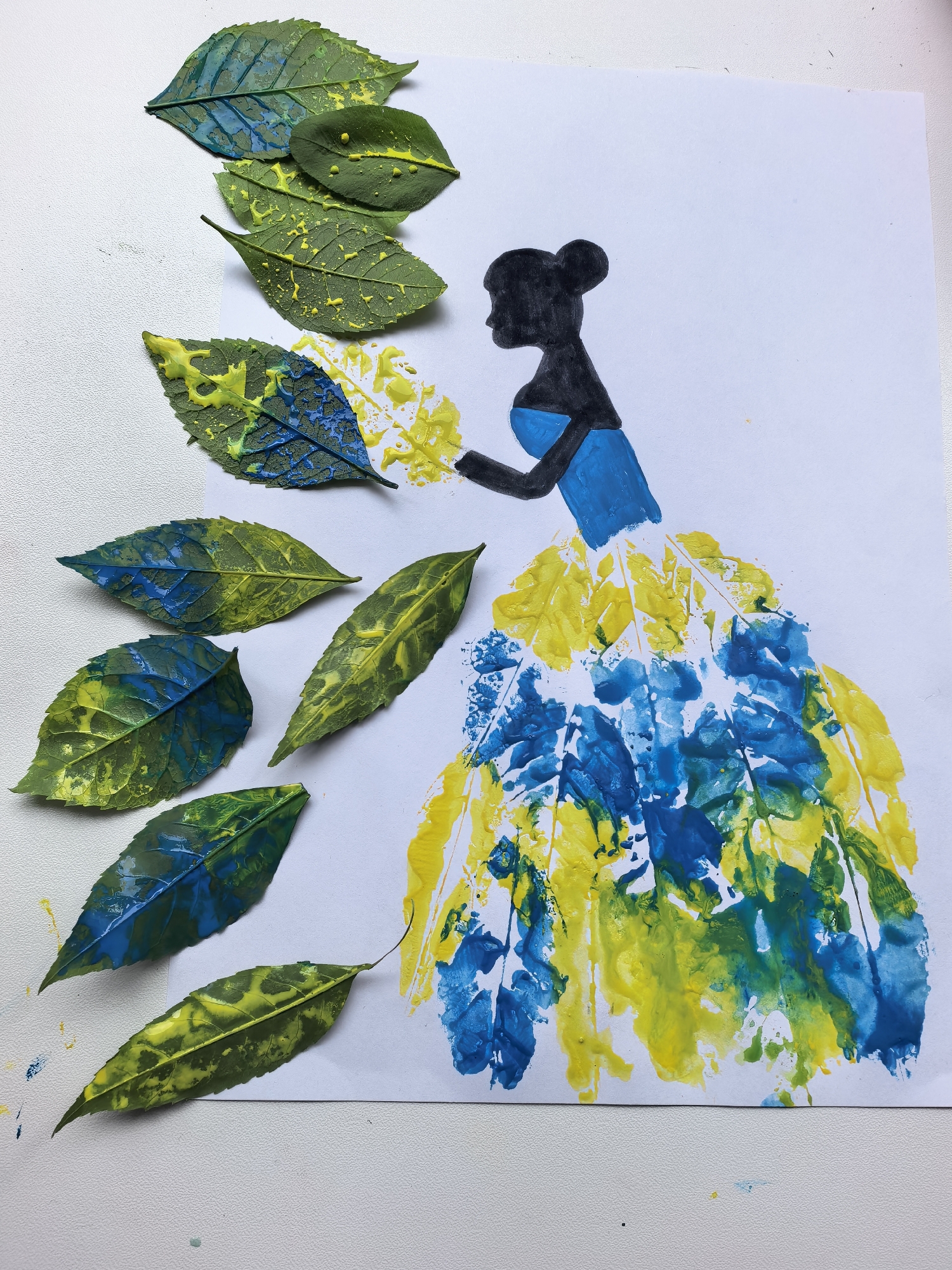 The image shows an art project created with leaves and paint. A silhouette of a woman, drawn in black, is depicted from the side. She is wearing a blue top and her hair is styled in a bun. Her dress appears to be made of leaf prints in blue and yellow paint, resembling a voluminous skirt. Several green leaves, some with yellow and blue paint splatters, are arranged on the left side of the paper, some overlapping with the printed dress. The background is a white surface, likely paper, with a textured white surface beneath it, possibly a table. There are no indications of the city of Sterlitamak, Russia, or any specific location within it.
