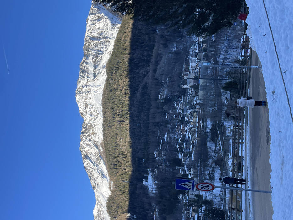 The image captures a panoramic view of a snow-covered alpine landscape, likely in Courmayeur, Italy. A majestic, rugged mountain, heavily dusted with snow and featuring rocky peaks, dominates the left side of the frame under a clear, bright blue sky. A faint airplane contrail is visible high in the sky.
Midway down the mountain, a dense, dark green forest of evergreen trees forms a prominent horizontal band, separating the sun-drenched upper slopes from the shadowed valley below. This band of trees casts a long, distinct shadow across the right side of the image, indicating either early morning or late afternoon.
In the mid-ground on the right, a charming town or village is nestled in the valley, partially obscured by the shadows. Its buildings, many with traditional pitched roofs, are also dusted with snow, giving the settlement a picturesque winter appearance.
In the immediate foreground, a paved road or street runs horizontally. To the right of the road, a snow-covered bank or path extends towards the edge of the frame. Two individuals are visible in this area. One person, wearing a light-colored puffer jacket and dark trousers, stands on the snow, facing away from the viewer and looking towards the town. Closer to the road, another person in dark clothing stands near a signpost.
The signpost displays two road signs: a blue square sign with a white triangle and a pedestrian symbol, and a white circular sign with a red border indicating a "30" speed limit. A brown guardrail or fence runs alongside the road. The overall scene conveys a serene winter day in a mountainous region, characterized by crisp air and dramatic natural beauty.