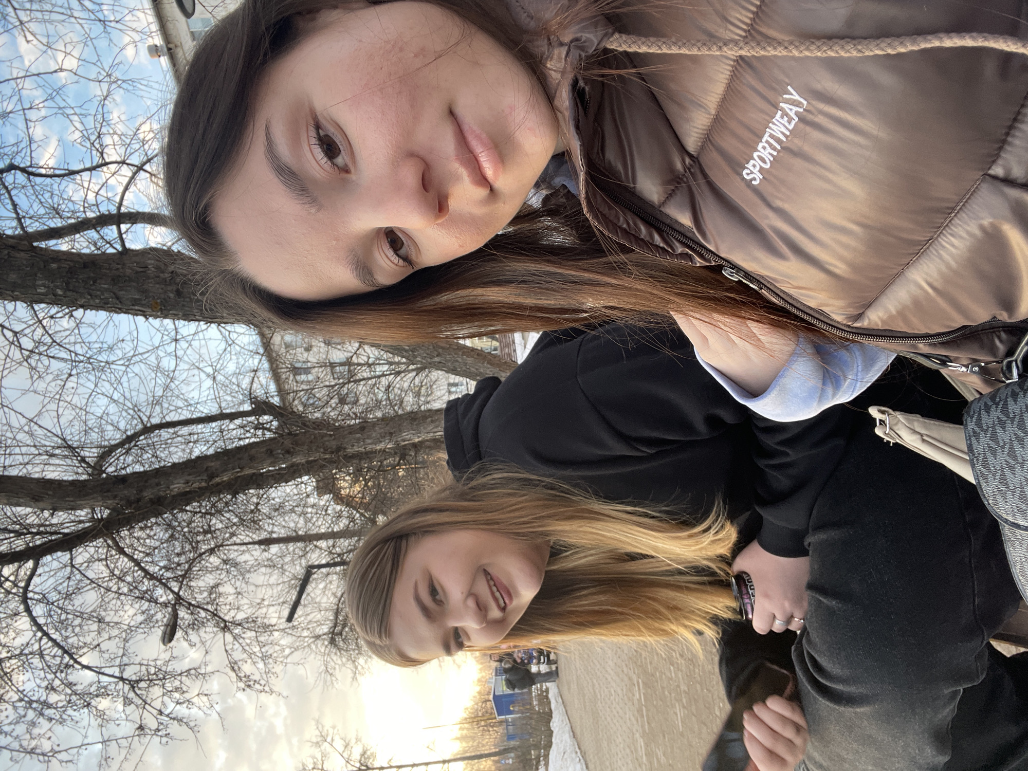 Two women are pictured in an outdoor setting. The woman at the top of the frame has dark hair and is wearing a brown puffer jacket with the word "SPORTWEAY" embroidered on the chest. The woman at the bottom of the frame has blonde hair and is wearing a black hoodie. They appear to be posing for a selfie or having their picture taken.
The background shows bare trees and a building partially visible at the top left. The lower portion of the image shows a sidewalk with some snow patches and what appears to be a fenced area with some structures in the distance. Based on the elements visible in the background, the photo may have been taken in a park or outdoor recreational area.
While I cannot specifically pinpoint the exact location within Chaikovsky, Russia without additional contextual clues, the visual cues of bare trees, possible snow, and urban elements suggest a park or public space in a city like Chaikovsky.